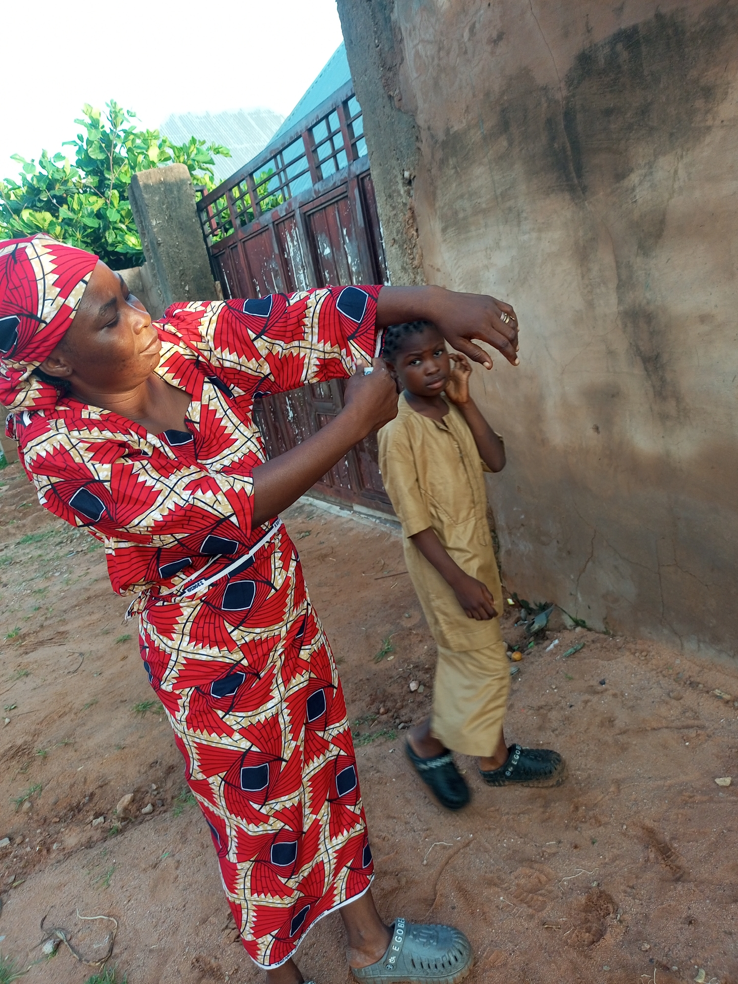 In Tempure, Nigeria, a woman in a vibrant red and yellow patterned dress and headwrap is tending to a young child. The woman is leaning down, her hands near the child's ear. The child, wearing a light brown outfit, stands facing the viewer with a slightly withdrawn expression and one hand touching their ear. Behind them, a textured, light brown wall dominates the background, with a rustic metal gate and some green foliage visible to the left. The ground is sandy and dusty, with scattered debris. The lighting suggests it is daytime, likely morning or late afternoon given the soft shadows. The overall scene appears to be outdoors, possibly in a residential area. The woman's gesture could indicate she is checking the child's ear or perhaps assisting them with something. The child's posture and expression convey a sense of gentle inquiry or slight discomfort. There is no visible text in the image.