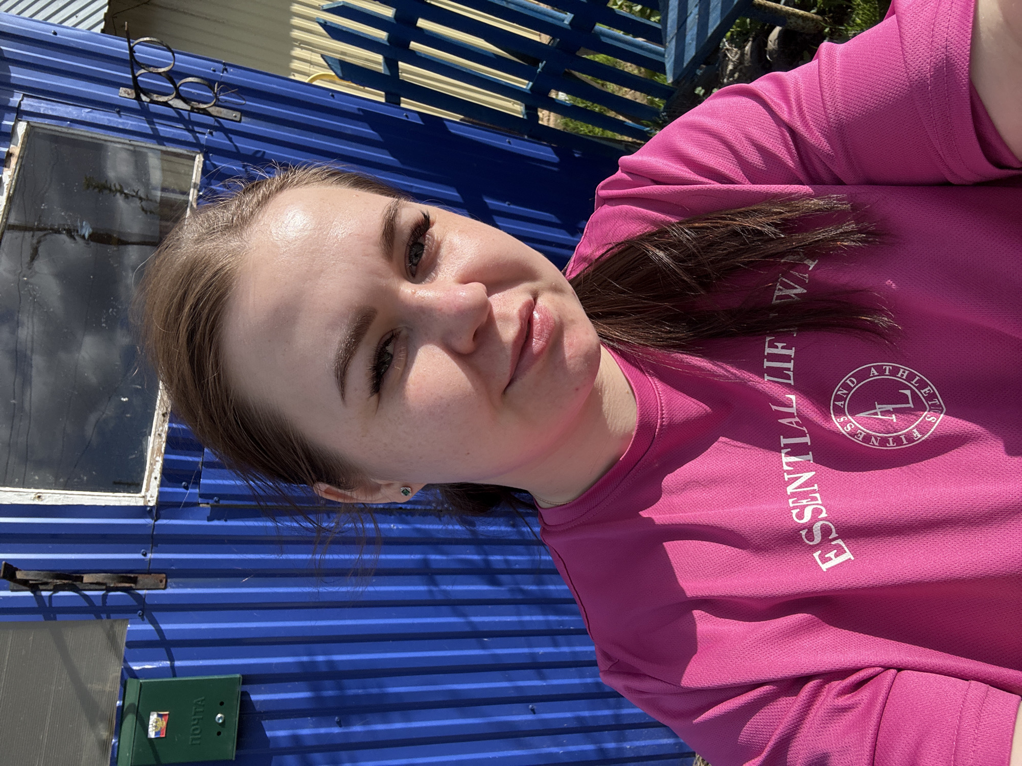 A woman is posing for a selfie outdoors in Ol'khovochka, Russia. She is wearing a pink athletic t-shirt with white text, and her hair is pulled back. Behind her is a corrugated blue metal structure with a window and a green mailbox. Sunlight illuminates the scene. A blue and white fence is partially visible in the background.