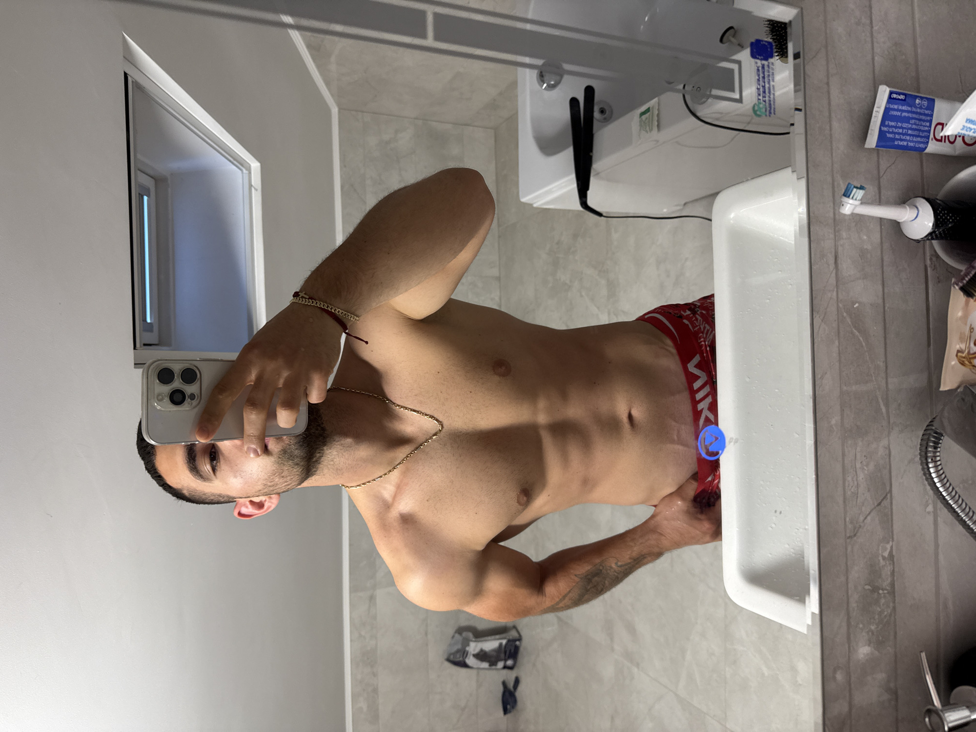 A shirtless man is taking a mirror selfie in a bathroom. He is holding a smartphone, obscuring part of his face. He is wearing red underwear with the word "NIK" visible on the waistband. A gold chain is around his neck and a bracelet is on his wrist. The bathroom contains a sink, toilet, shower, and window. Toothpaste and a toothbrush are visible on the counter. There is no indication that this image was taken in the city of Gabrovo, Bulgaria.