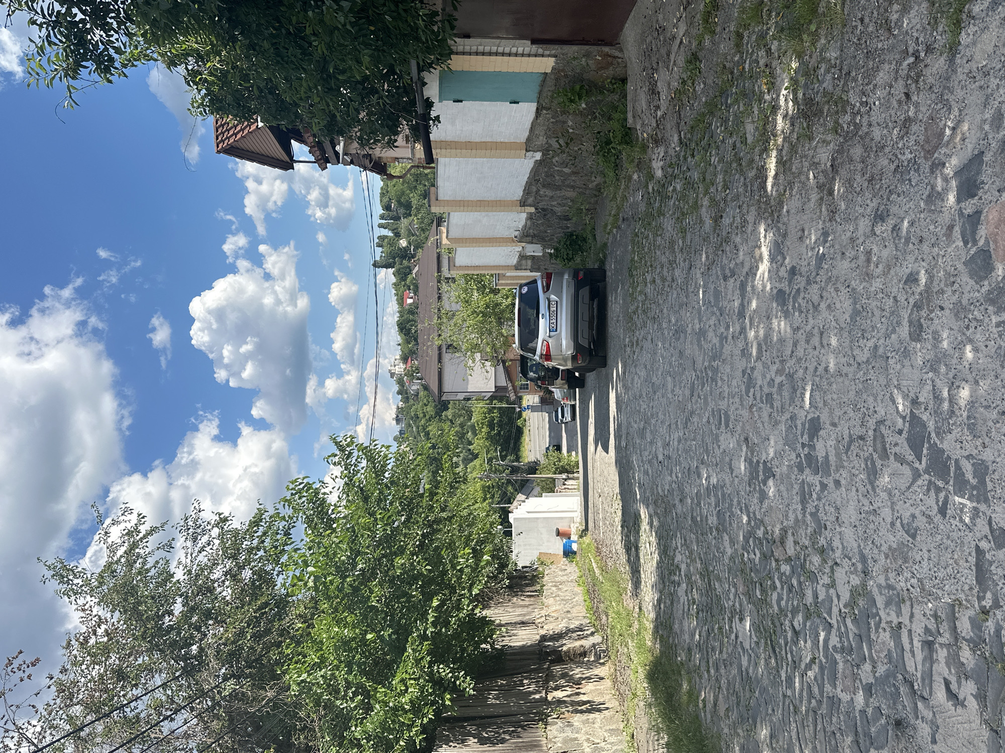 The image depicts a street scene in Kyiv, Ukraine. The perspective is from the side of the street, looking down a cobblestone pathway towards a silver SUV parked on the right side of the image. The SUV has the license plate "KA 5506 IC". Behind the SUV, there are several garage doors, some painted white and one a teal color, against a stone wall.
To the left of the street, a wooden fence runs along the property line, with lush green trees and foliage growing over it. Further in the background, a landscape of houses and trees is visible, with a hill in the distance. The sky above is a vibrant blue, dotted with large, fluffy white clouds, suggesting a sunny day with pleasant weather. The sunlight casts shadows on the cobblestone street, indicating it is likely daytime.
There are no people clearly visible and engaged in any specific activity, though a blurred figure can be seen near the garages. The overall impression is a quiet residential street in an area with a mix of older architecture and natural greenery.