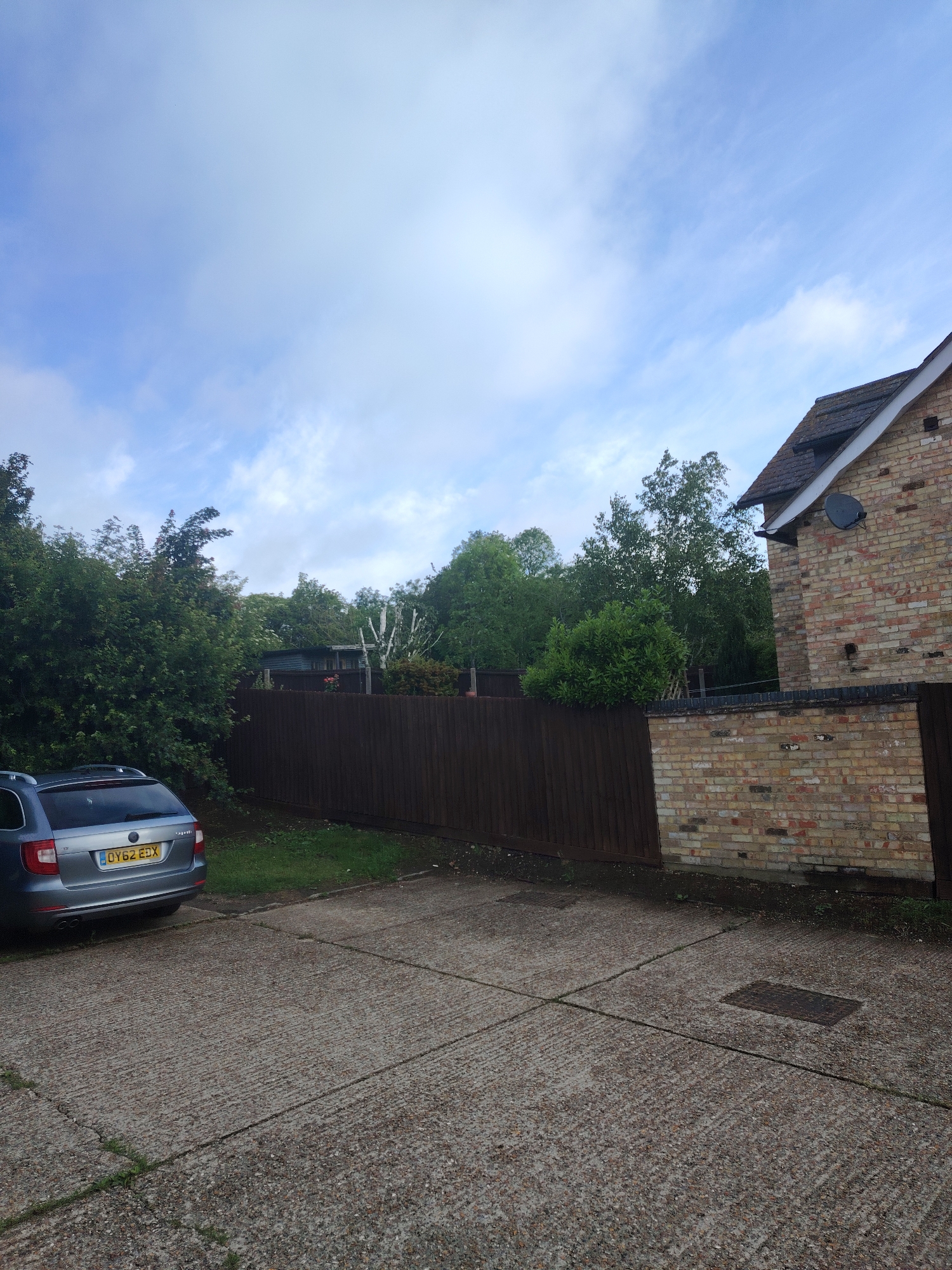 The image presents an outdoor residential scene in Huntingdonshire, United Kingdom, under a partly cloudy sky. The foreground consists of a concrete driveway or parking surface, characterized by textured parallel grooves and visible expansion joints or cracks, with some minor weed growth. A rectangular metal manhole cover is embedded in the concrete on the right side.
On the left, the rear section of a silver-grey estate car, featuring roof rails, is parked, displaying the UK registration plate 'OY62 EDX'. Behind the car and along the left-midground, a dense collection of green foliage, comprising various trees and shrubs, forms a natural barrier.
A dark brown wooden panel fence stretches horizontally across the mid-ground, demarcating the concrete area from a grassy strip. Beyond this fence, a small shed or outbuilding is partially visible amidst a grove of trees, which includes one with distinctively light-colored bark.
To the right, a low wall constructed from varied yellow and light brown bricks continues the boundary, transitioning into the side of a two-story residential building made of the same multi-toned bricks. The house features a dark grey tiled roof, incorporating a visible dormer or roof extension, and has a black satellite dish mounted on its exterior brick wall. No individuals are present in the depicted scene.