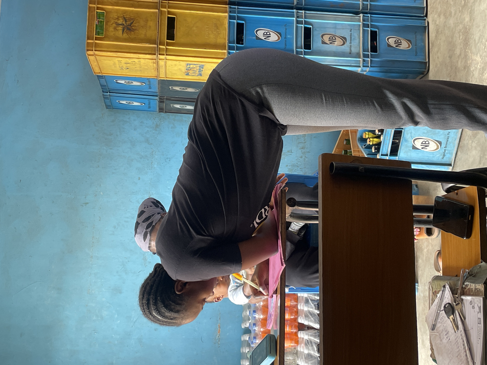 The image shows a woman in what appears to be a small shop or store. She is wearing a black top and gray pants, leaning over a table or counter, and is likely writing or working with something on the table. She has braided hair and is wearing a head covering. On the table, there are bottles of water or drinks and papers. Behind her, there are stacks of plastic crates, some yellow and some blue, with the "NB Nigerian Breweries Plc" logo, suggesting this is a shop that sells beverages. The wall is painted a bright blue. The presence of the crates and the beverages suggest a commercial setting, possibly a small retail shop or kiosk, typical of small businesses in Jos, Nigeria. There's also what appears to be a telephone on the table.