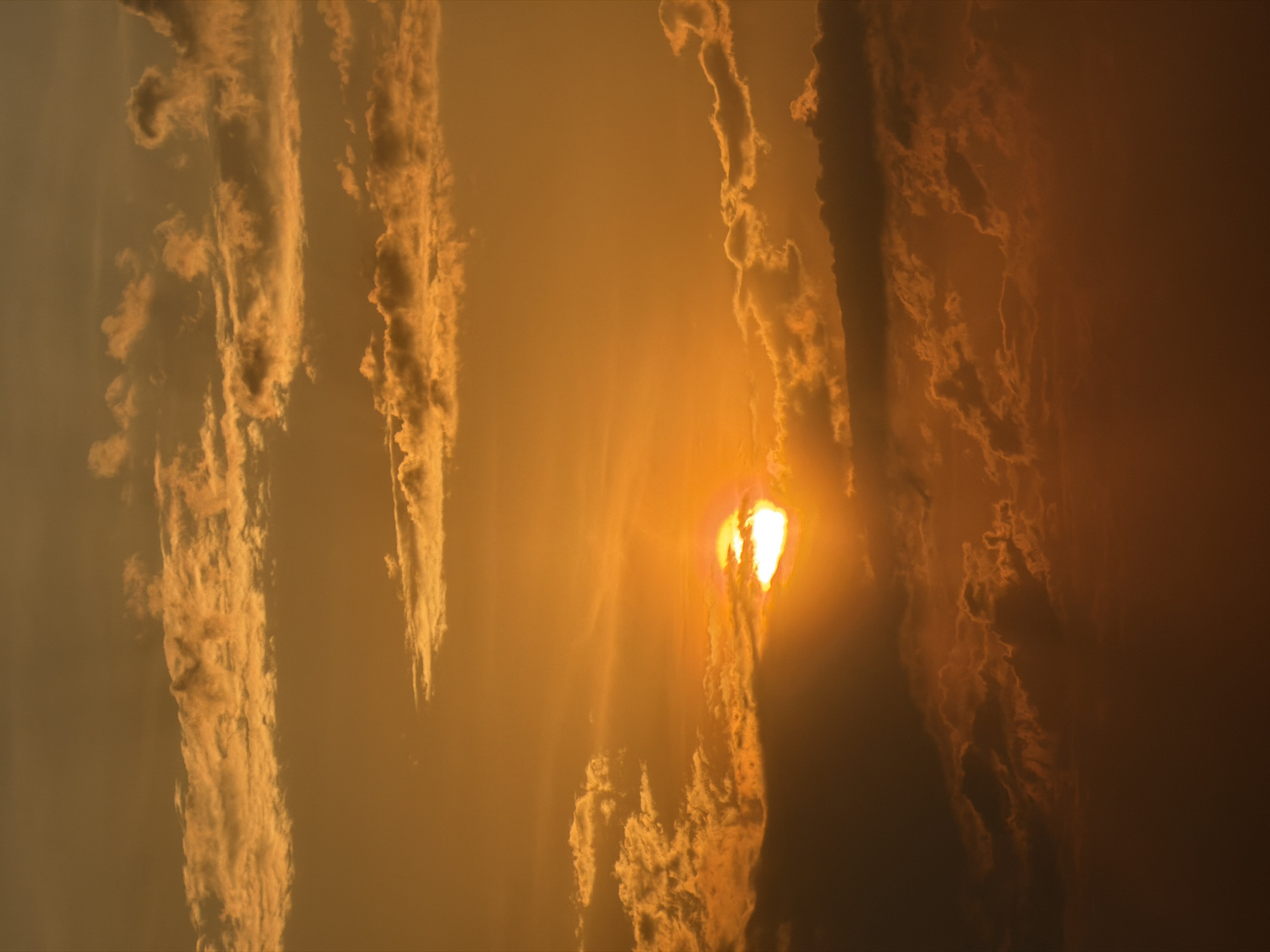 The image depicts a sunset scene in an unspecified location. The sky is filled with clouds, varying in density and illuminated by the setting sun, which is positioned in the center of the frame. The sun's light permeates the clouds, creating a warm, orange and yellow color cast across the scene. The overall atmosphere is hazy and the clouds appear to be vertically oriented. There are no people or distinct objects visible other than clouds and sunlight.