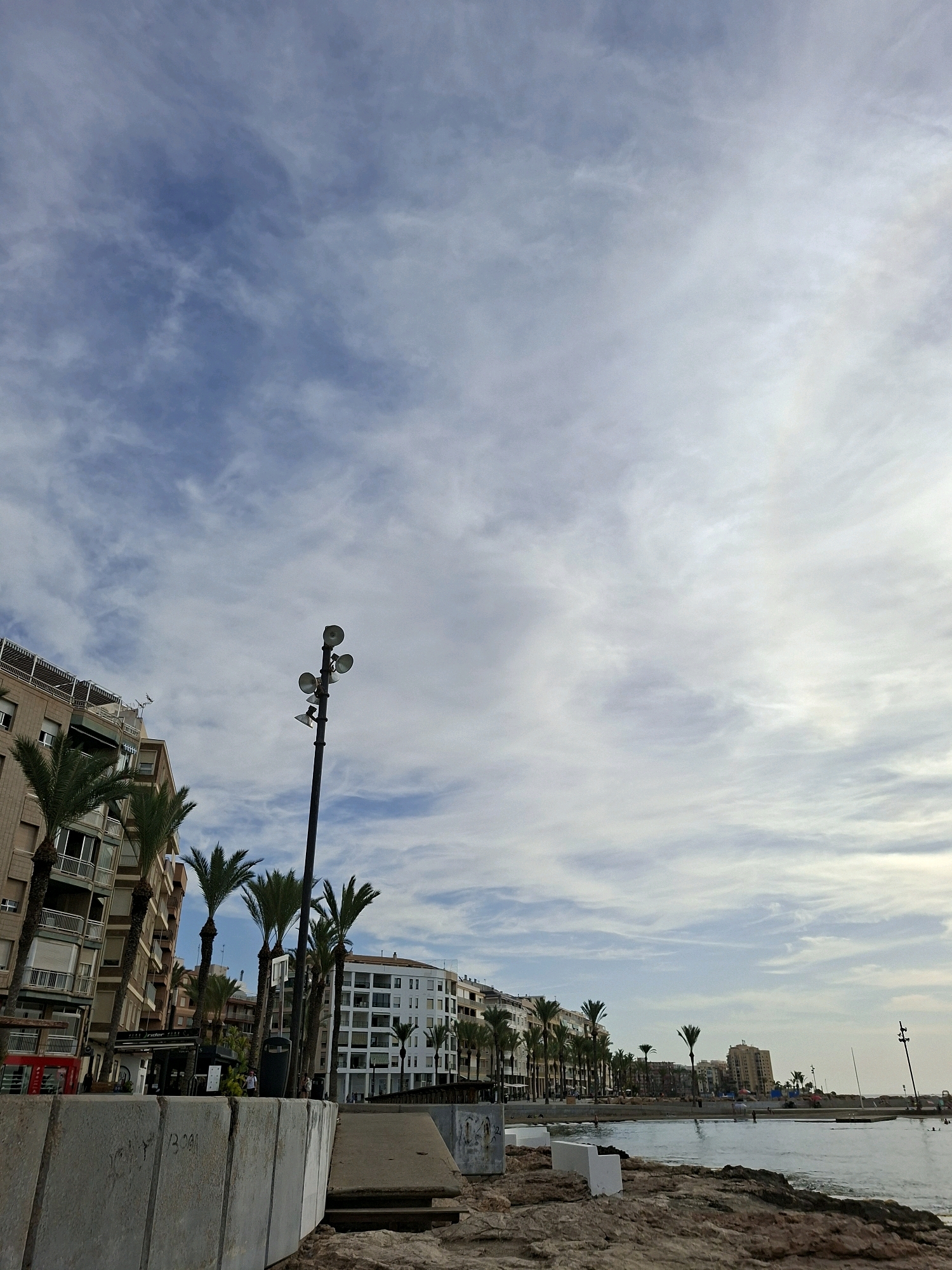 The image captures a view of the waterfront in Torrevieja, Spain, with the Mediterranean Sea on the right and a row of buildings and palm trees on the left. In the foreground, there are large concrete blocks, suggesting a pedestrian walkway or barrier. The sea is calm, with some light ripples. A few people can be seen near the water's edge and on the distant promenade. The buildings are a mix of residential and commercial, with balconies and awnings visible. Palm trees are scattered along the promenade, adding to the coastal atmosphere. The sky is partly cloudy, with a mix of blue sky and white clouds. A streetlamp with multiple lights is prominent on the left side of the image, silhouetted against the sky. In the far distance, more buildings and a boat are visible on the horizon. The overall scene depicts a typical day at the seaside in a Spanish coastal city.
