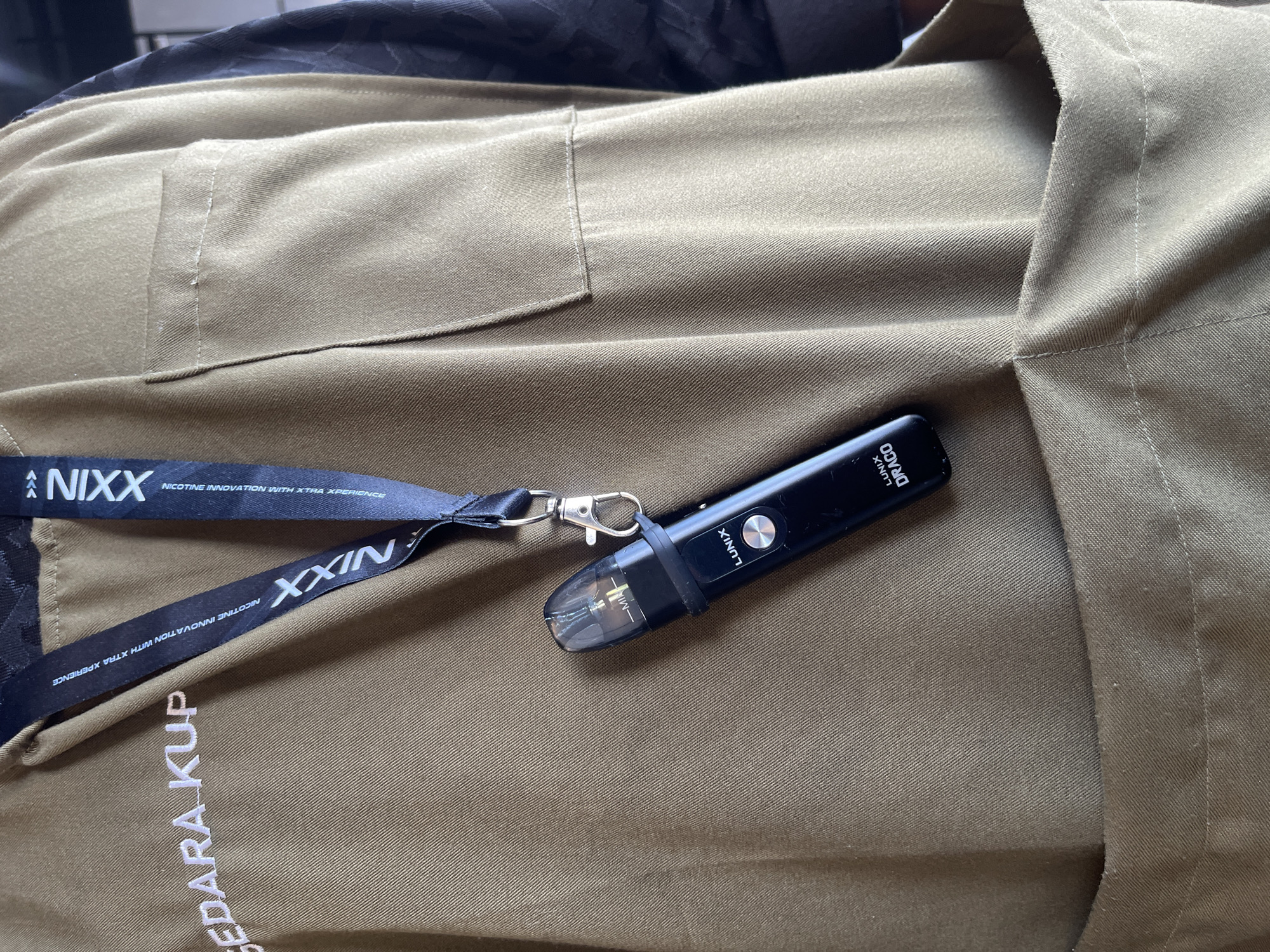 The image shows a close-up of a person's torso, wearing a brown garment with embroidered text on the left side that reads "SEDARA KUP". A black lanyard with white text "NIXX NICOTINE INNOVATION WITH XTRA XPERIENCE" is visible around the neck and draped over the garment. A black vape device, branded "LUNIX DRACO", is attached to the lanyard and hangs near the chest area. The garment has a breast pocket on the right side. The scene is indoors, with the background suggesting a neutral setting. There is no visual information to confirm the location as Bireuenmeunasahdayah, Indonesia.