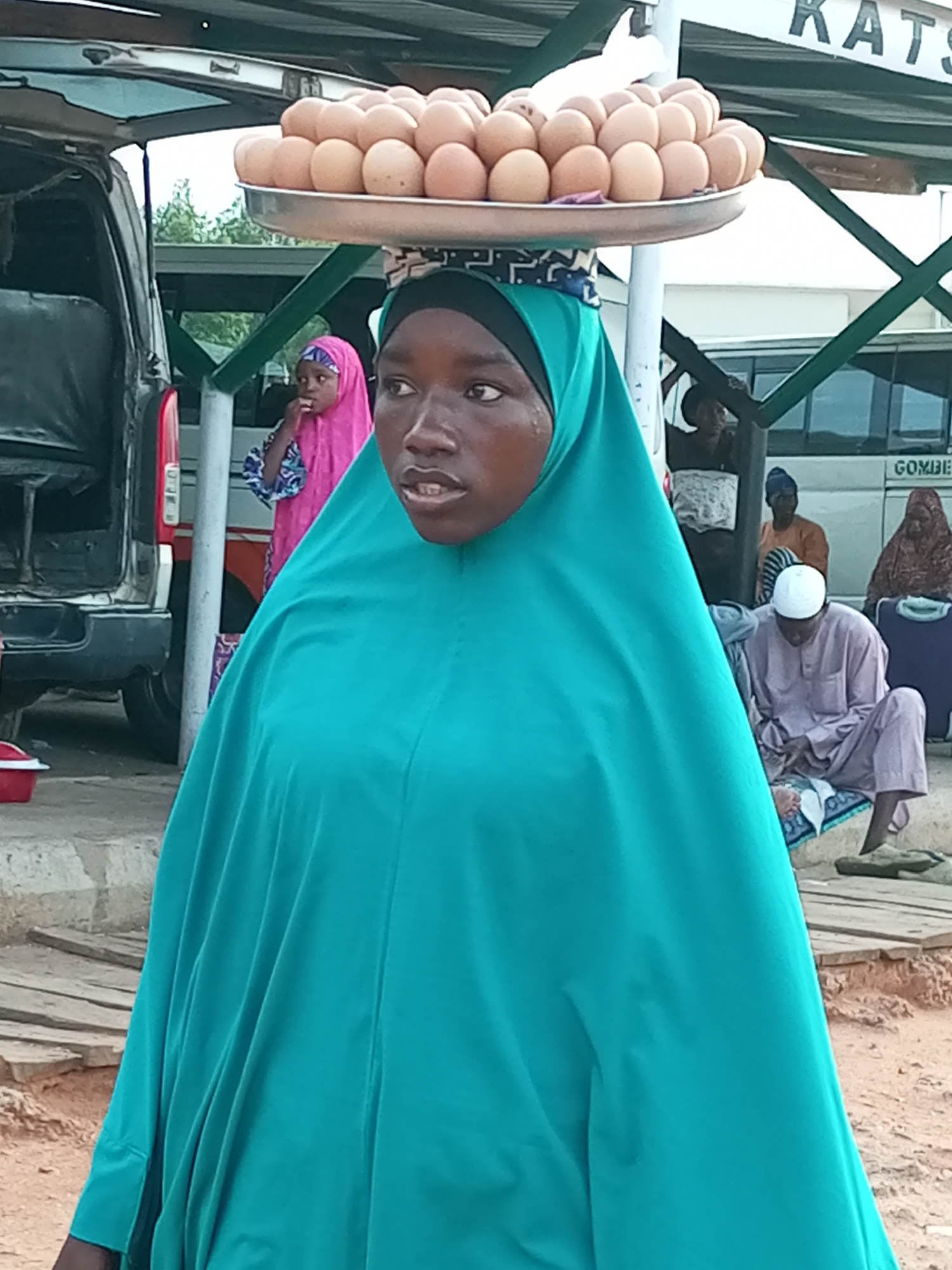 A young Nigerian woman, wearing a bright turquoise hijab and garment, is depicted in the foreground of this image, with a tray of eggs balanced on her head. Her gaze is directed to her right, and her mouth is slightly open, suggesting she may be speaking or in the process of doing so. Behind her, other individuals are visible, including a young girl in a pink hijab and a man in a white cap and light purple clothing, seated on a mat. The scene appears to be an outdoor market or roadside stall in the city of Gombe, Nigeria, with a vehicle parked to the left and what looks like a bus in the background, possibly indicating a transportation hub or busy thoroughfare. The ground is dusty and uneven, with wooden planks suggesting a temporary or improvised walkway. The overall atmosphere suggests a moment of daily commerce or travel in a bustling urban environment.
