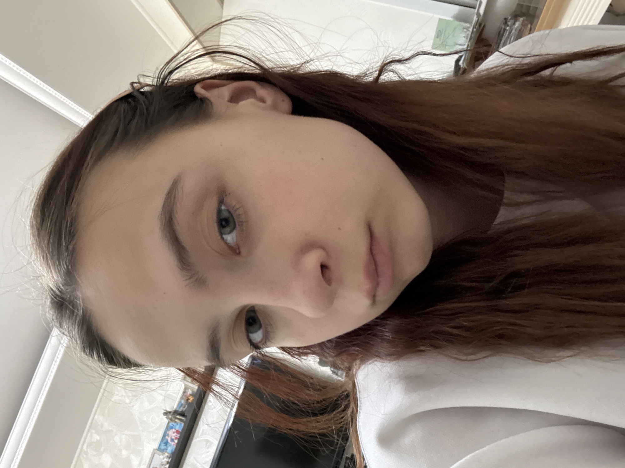 A close-up, slightly angled headshot of a young woman. Her face is the primary focus, displaying fair skin, light-colored eyes, and brown hair with reddish tones. She is wearing a white garment. The background is blurry and indistinct, but suggests an interior setting with light-colored walls and possibly a TV. The location in the city of Berezniki, Russia, cannot be confirmed based on the visual data provided.