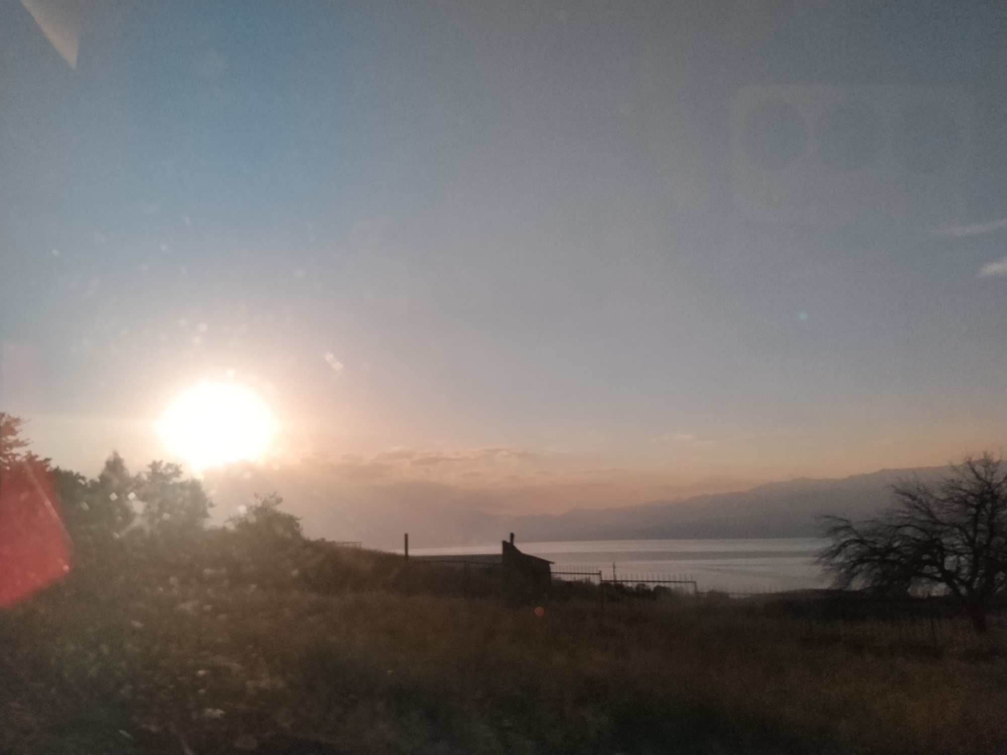 The image depicts a sunset or sunrise over a body of water, likely a lake, with a mountainous landscape in the distance. The foreground is filled with dry grass and some trees, one of which is bare and silhouetted against the sky. A small building with a chimney is visible near the water's edge, surrounded by a fence. The sky is a gradient of pale blue to orange and pink hues, with the sun a bright, blurry orb. There are no people or animals visible in the scene. The overall impression is one of a tranquil, natural landscape at twilight. The location is identified as Chkalov, Kyrgyzstan.