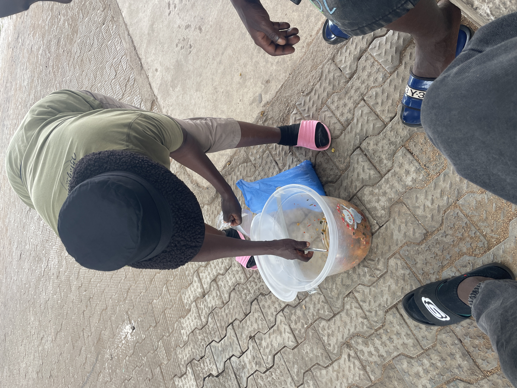 This overhead shot, taken in Jos, Nigeria, captures an outdoor scene featuring at least two individuals on a paved surface during daytime.
The primary subject, positioned towards the left, is seen bending over a large, clear plastic container, seemingly stirring or serving food with a metal spoon. This person wears a green t-shirt, light-colored shorts, and a black fuzzy bucket hat. On their right foot, they have a black sock and a bright pink slide sandal. Their left hand appears to be holding a transparent plastic bag, possibly for packaging the food. The large container holds a reddish-orange food mixture with visible pieces, suggestive of a stew, rice dish, or salad, and features cartoon-like holiday decals (a Santa Claus and reindeer) on its side. A blue plastic bag lies on the ground next to the container.
Other partially visible individuals surround the central activity. To the top right, a person's dark-skinned hand is visible, along with their legs and feet, wearing dark jeans and blue slide sandals with "Y-3" written in white. To the bottom right, another person's foot, clad in a black slide sandal with a white circular logo (resembling an 'O' or an eye shape), is seen wearing dark jeans.
The ground consists of grey interlocking paving stones with some scattered dirt and debris, alongside a smoother, lighter concrete area. The bright lighting suggests it is day, and the casual attire and outdoor setting are consistent with street vending or a community gathering. No specific emotions are discernible from this vantage point, but the scene depicts an ordinary, functional interaction.