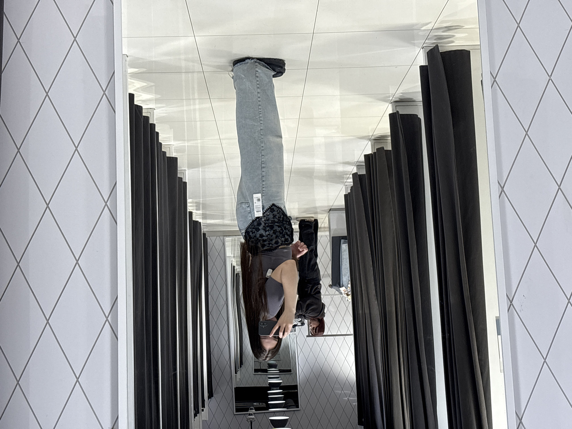 A young woman is reflected in a mirror inside a dressing room. The dressing room has white, diamond-patterned tile walls and black curtains. The woman is wearing a gray shirt, blue jeans, and black shoes. She is holding a phone and taking a picture of herself in the mirror. A second woman, wearing a black hoodie and black pants is also visible in the reflection. The location is potentially a retail establishment, possibly in Yekaterinburg, Russia.