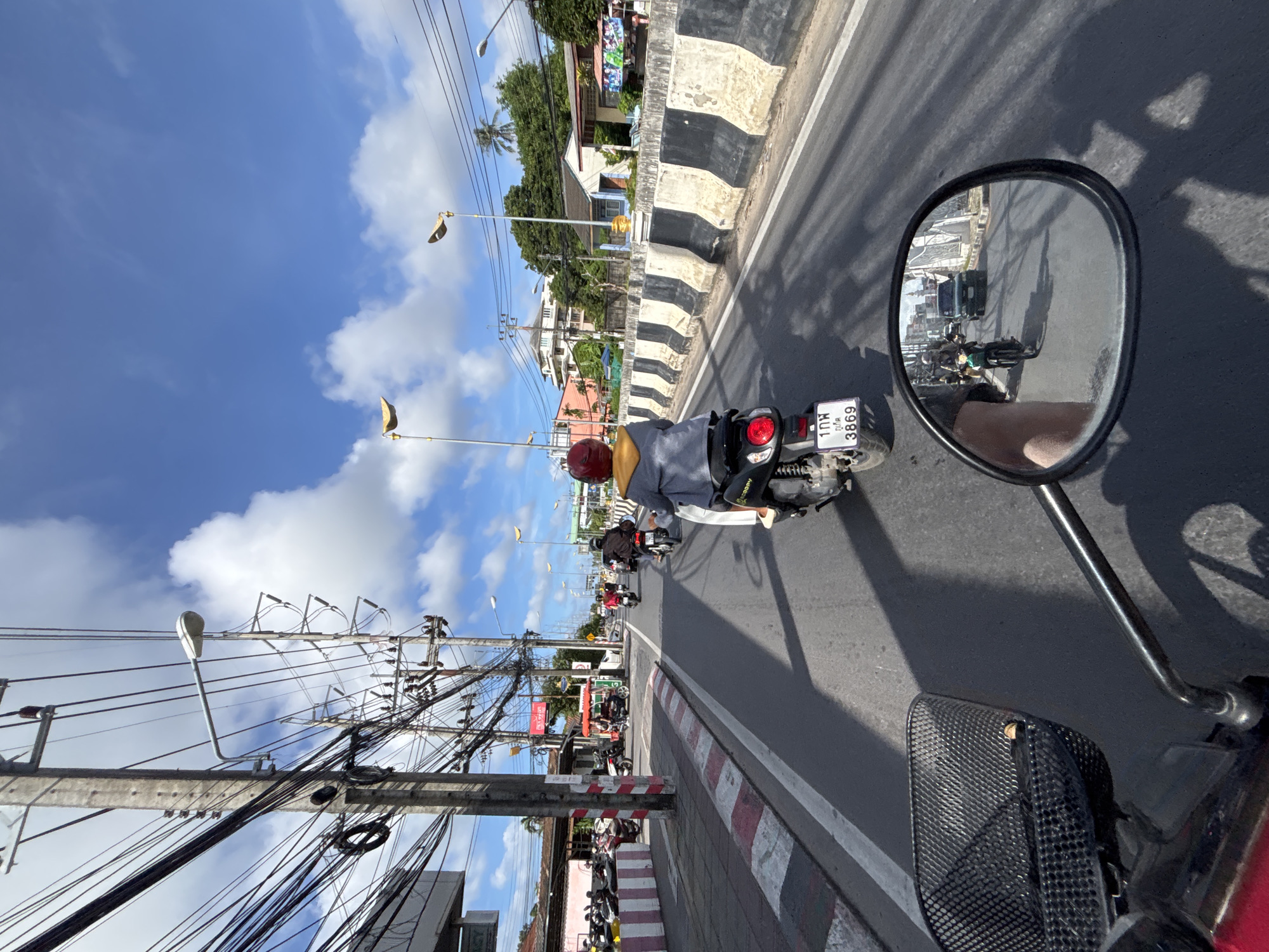 This is a point-of-view shot from a motorcycle traveling on a street in Rawai, Thailand. The road is asphalt, with a white line marking the lane. To the left is a sidewalk with a red and white striped curb, and a row of parked motorcycles. Further left, buildings are visible, including one with a red awning. Overhead, numerous power lines and poles crisscross the sky. Streetlights are mounted on poles along the road. On the right side of the road, a concrete barrier with black and white diagonal stripes separates the road from an area with buildings and trees. In the foreground, the motorcycle's handlebars, a black basket, and a rearview mirror are visible. The rearview mirror reflects other motorcycles and a car on the road ahead. The sky is blue with scattered white clouds. A person on a motorcycle in front of the viewer is wearing a gray jacket and a red helmet. Several other motorcycles are also on the road, some carrying riders. The license plate of the motorcycle directly in front reads "1กพ 3869".