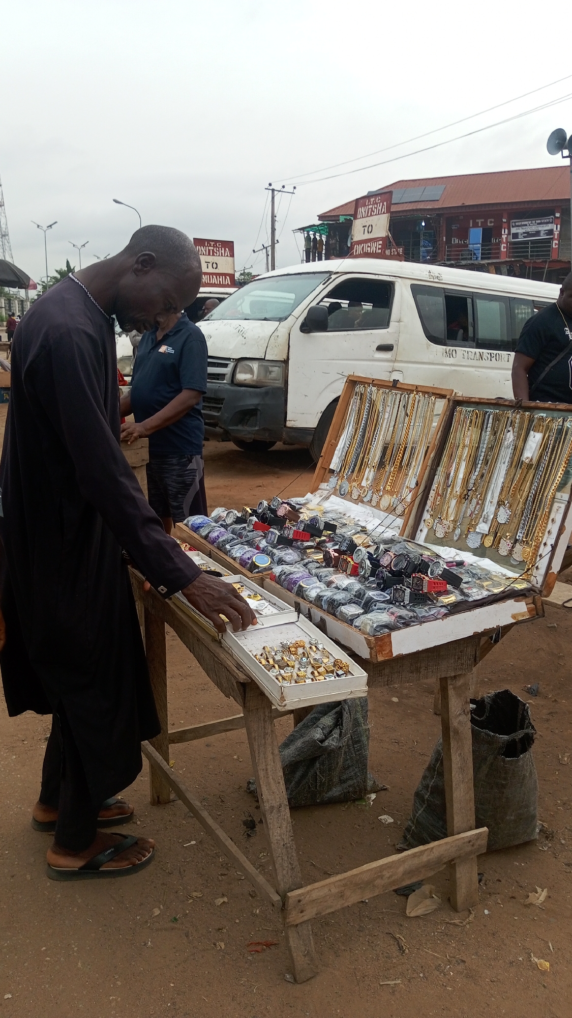 An outdoor market scene depicts a man in a dark kaftan and sandals inspecting jewelry at a roadside stall. He leans over a simple wooden table, his hand reaching towards a white tray filled with numerous gold-colored rings. To his right, two larger display cases are open, showcasing a wide array of merchandise: one is packed with various watches, many still in plastic packaging, and the other features rows of gold-colored necklaces with pendants.
In the background, a white passenger van, marked "IMO TRANSPORT" on its side, is parked, suggesting a transport hub or busy street. Another man in a blue polo shirt and patterned shorts stands near the van. The ground is reddish-brown earth, indicative of an unpaved or dirt area.
The sky is overcast, suggesting daytime with muted lighting. In the distance, several signs are visible. One prominent sign reads "I.T.C ONITSHA TO MUAHIA," and another, partially obscured, indicates "I.T.C ONITSHA TO OKIGWE." A building with a red roof and solar panels is also visible in the background, bearing the "I.T.C" text. Utility poles and an antenna tower further emphasize an urban or semi-urban setting. Two coarse, dark sacks rest on the ground beneath the jewelry table. The scene captures a typical bustling market activity in a Nigerian town like Onitsha.