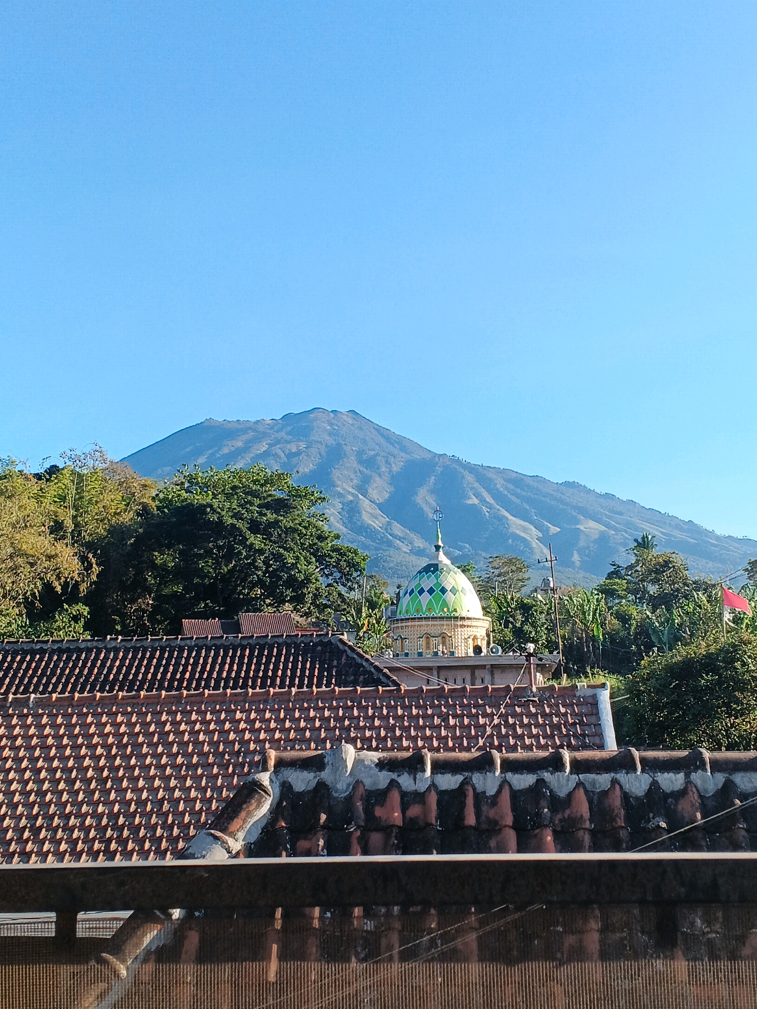 The image depicts a serene landscape with a prominent mountain in the background under a clear blue sky. In the foreground, several tiled roofs of houses are visible, with the closest one showing an aged, textured surface. Nestled between the houses and the mountain is a mosque with a distinct green and white patterned dome, topped with a crescent moon and spire. Lush green trees and foliage surround the mosque and dot the landscape leading up to the mountain. A red flag is seen fluttering on the right side of the image. The scene appears to be a peaceful view of a village or town, likely Bocek, Indonesia, with the mountain serving as a majestic backdrop.