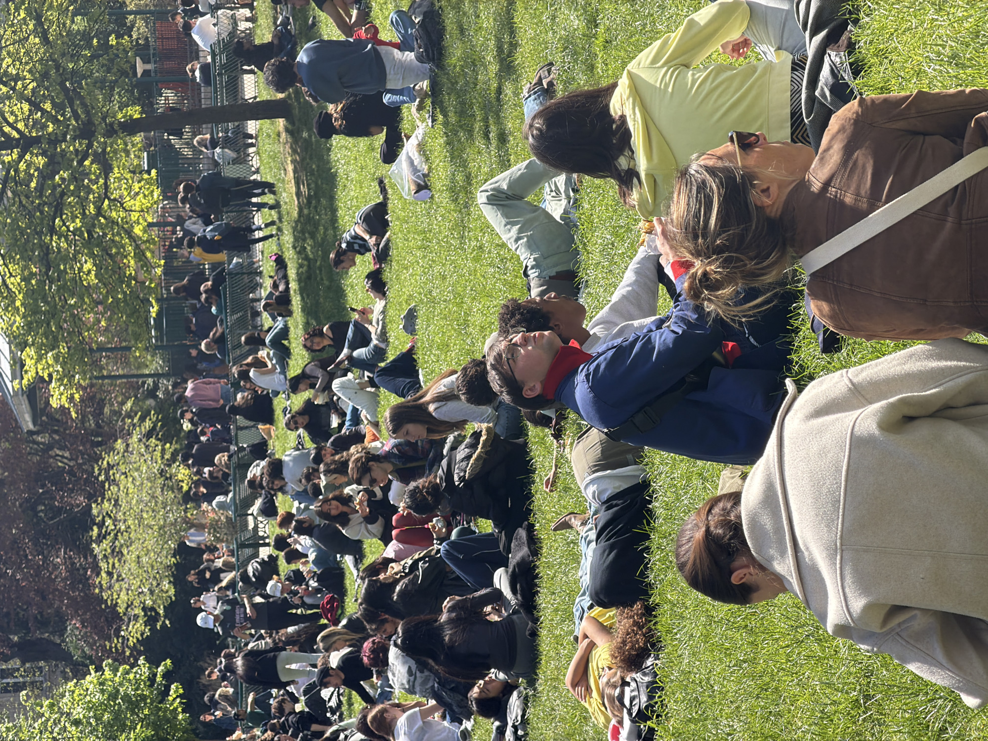 A large group of people is lying on the grass in a park, seemingly facing the same direction. Most of them are young adults, possibly students. The environment appears to be a grassy area within a park setting. There are trees in the vicinity. The location is Paris, France. In the background, there's a fence and some buildings, possibly indicating a more urban setting. People are mostly lying down, some are talking and engaging with others nearby. Some people are wearing coats and sweaters indicating that the weather is not very warm.