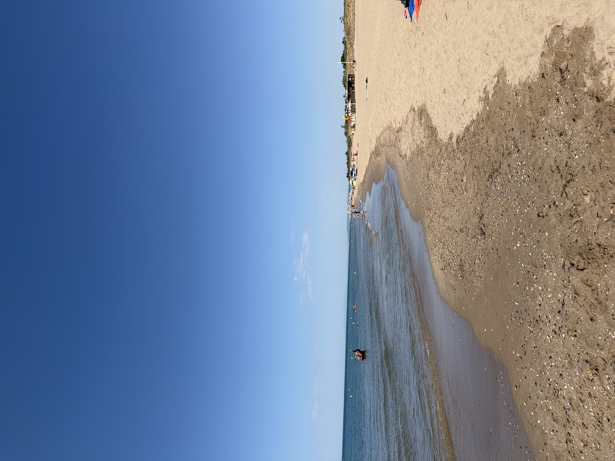 The image shows a view of the beach in Sérignan, France, on a sunny day. The sky is clear and blue. The sea is calm and appears to be a deep blue. Along the shoreline, there is a sandy beach with some scattered shells and debris. In the water, several people are visible swimming or wading. Further down the beach, there are some beach umbrellas, a few buildings, and more people relaxing on the sand. The overall scene depicts a typical summer day at a popular seaside resort.