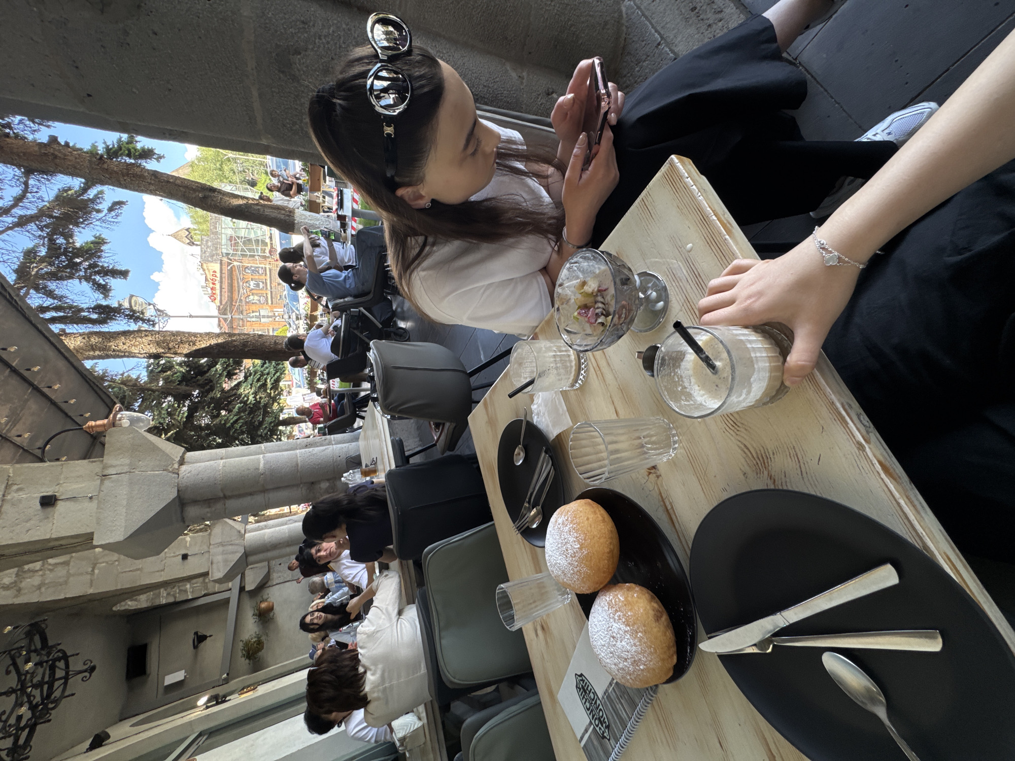 The image shows an outdoor cafe setting in Gyumri, Armenia. In the foreground, on a wooden table, are two powdered donuts on a black plate, a black oval plate with a knife, fork, and spoon, two empty glasses, and a glass of iced coffee with a straw. A woman in a white t-shirt and black pants is seated at the table, looking at her phone. She is wearing large sunglasses on her head. In the background, other patrons are seated at tables, and trees and buildings are visible. The architecture in the background suggests a historical city.