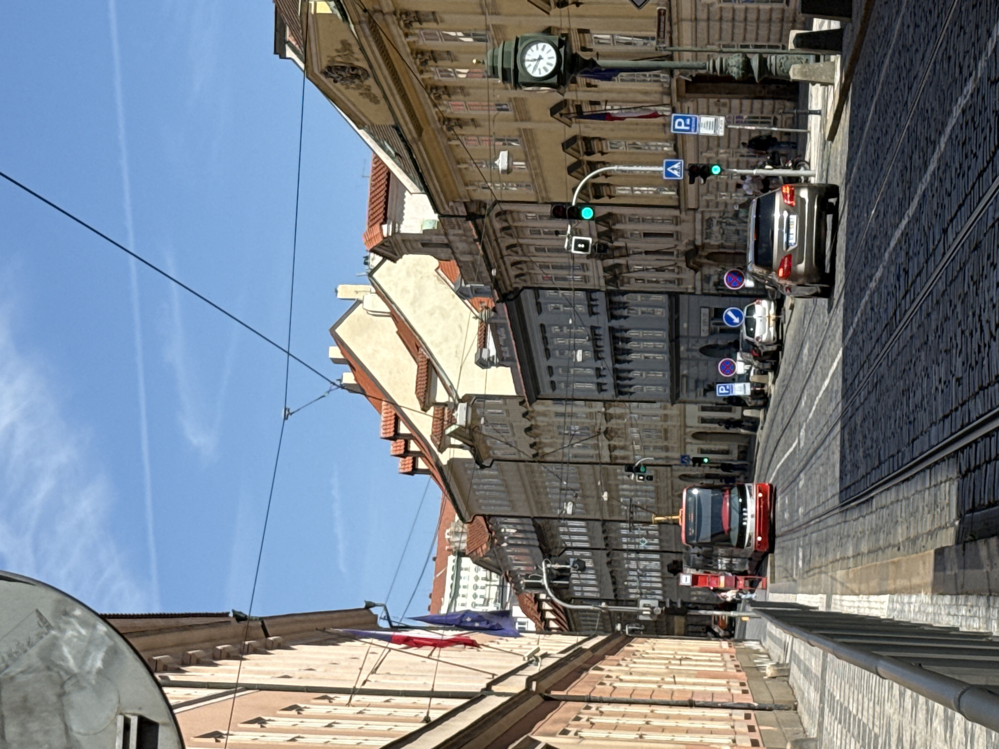 The image depicts a street scene in Prague, Czechia, likely in the historic Old Town or Lesser Town given the architecture. In the foreground, a red and white tram is stopped on tram tracks laid into a cobblestone street. Several cars are visible on the street, including a silver Mercedes-Benz sedan in the middle ground. The buildings lining the street are characteristic of Prague's historical architecture, with multiple stories, ornate facades, and red-tiled roofs. Flags, including the Czech flag and what appears to be the European Union flag, are visible on one of the buildings. Streetlights, traffic signals (one showing green), and street signs are present. The sky is blue with a few wispy clouds. The overall impression is a sunny day in a busy urban environment.