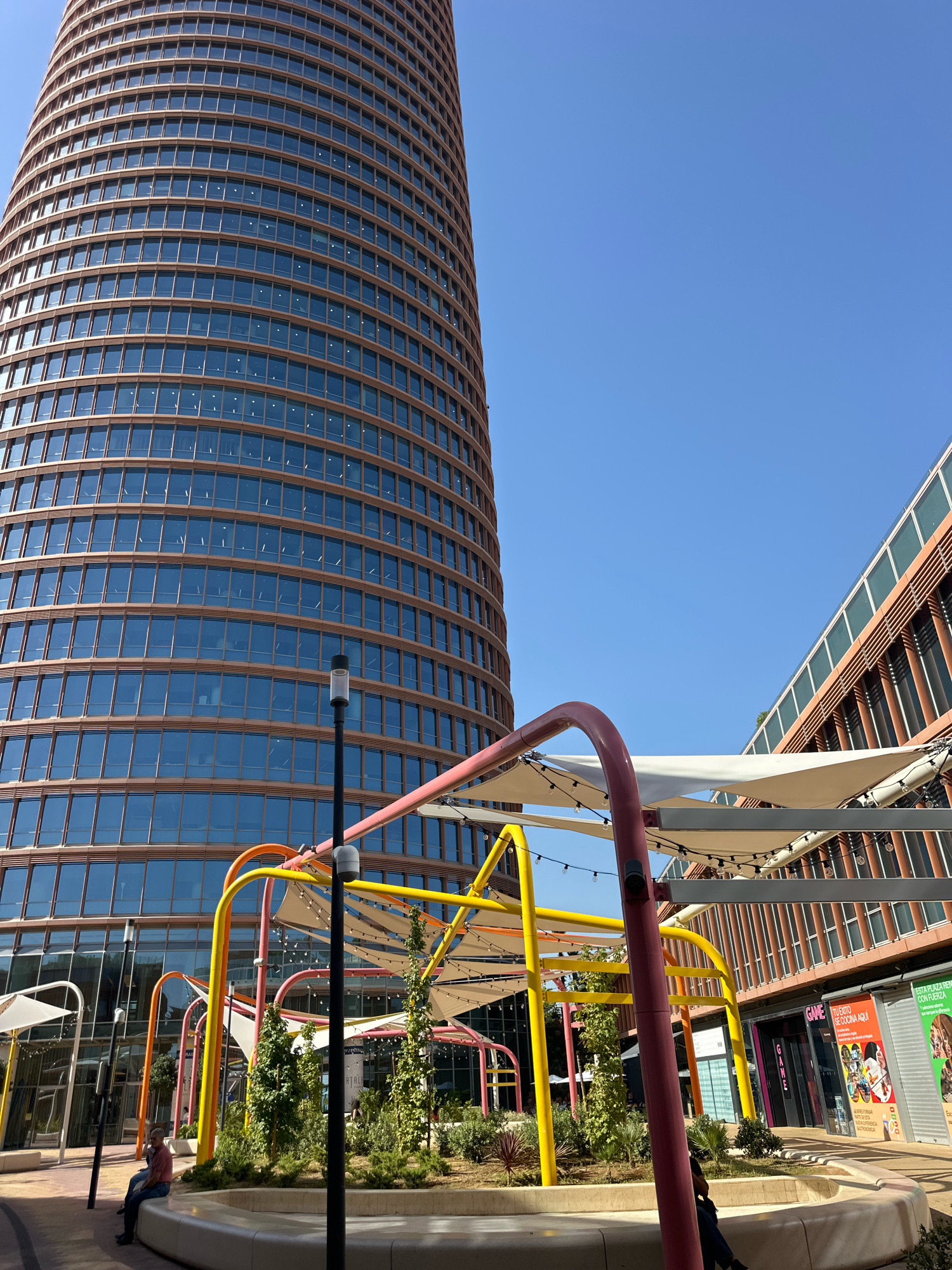 The image captures an outdoor area in Seville, Spain, with the prominent Torre Sevilla skyscraper dominating the left side of the frame. The skyscraper features a curved, glass-covered facade in a warm brown hue, with numerous horizontal window panes. To the right of the skyscraper, another modern building with a similar brown exterior and vertical window elements is visible, angled towards the sky. In the foreground, a vibrantly colored, abstract playground structure with red, yellow, and orange arches is set against a backdrop of greenery and a light-colored curved bench. String lights are draped across the playground's canopies. A lamppost stands to the right of the playground. A single person is seated on the bench in the lower left quadrant of the image, facing away from the camera. The sky is a clear, bright blue. A sign on the building to the right reads "GAME TU EXITO SE COCINA AQUI".