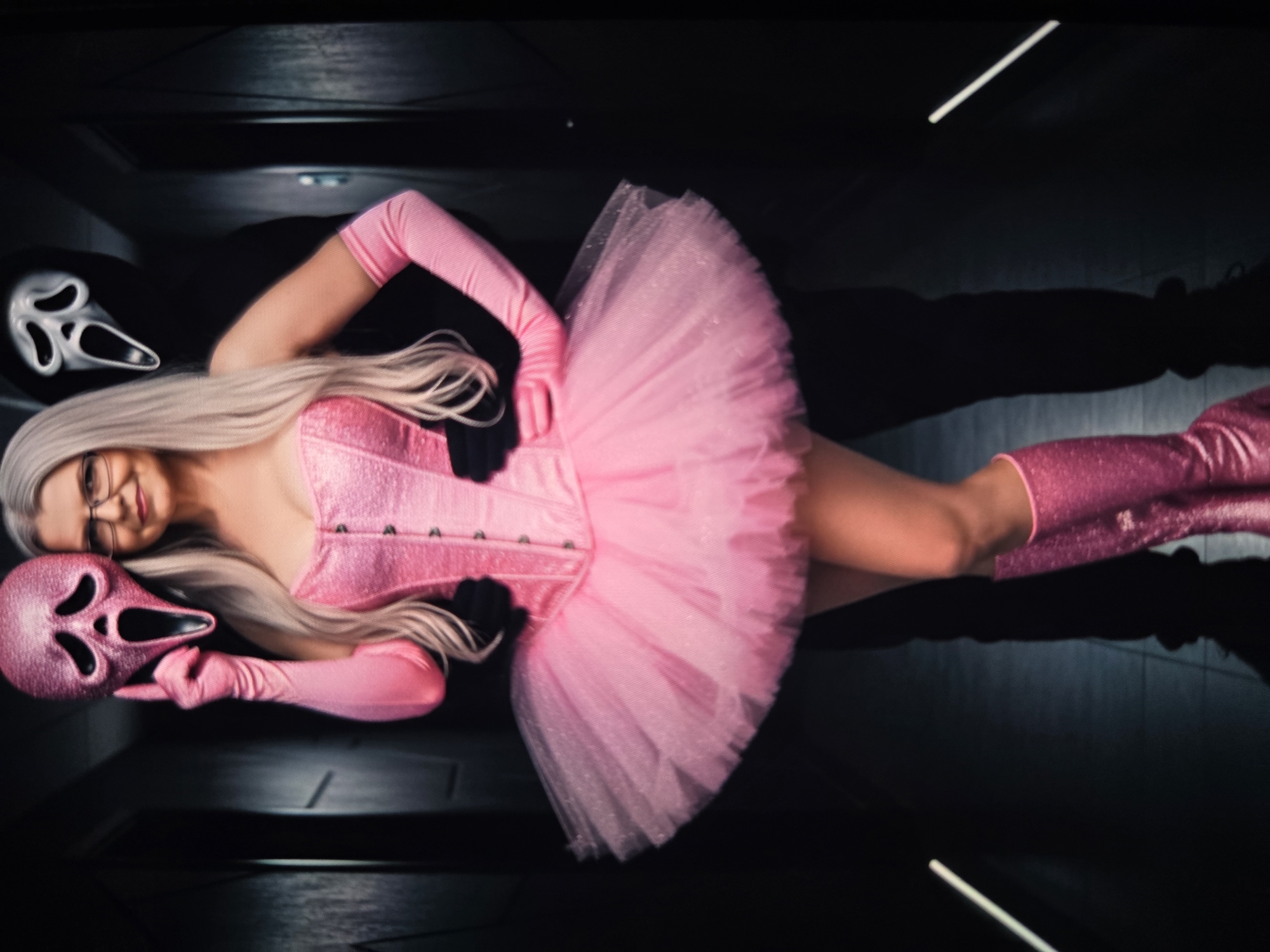 In a dimly lit hallway, a woman with long, platinum blonde hair and glasses stands in the foreground, holding a pink sequined mask with a gaping mouth to her face. She wears a matching pink sequined corset and a voluminous pink tulle tutu. Pink sequined gloves extend up her arms, and she is also wearing knee-high pink sequined boots. Behind her, a figure in black, wearing a white Ghostface mask from the Scream franchise, stands partially obscured. The hallway has dark, textured walls and a reflective floor with faint linear light sources.