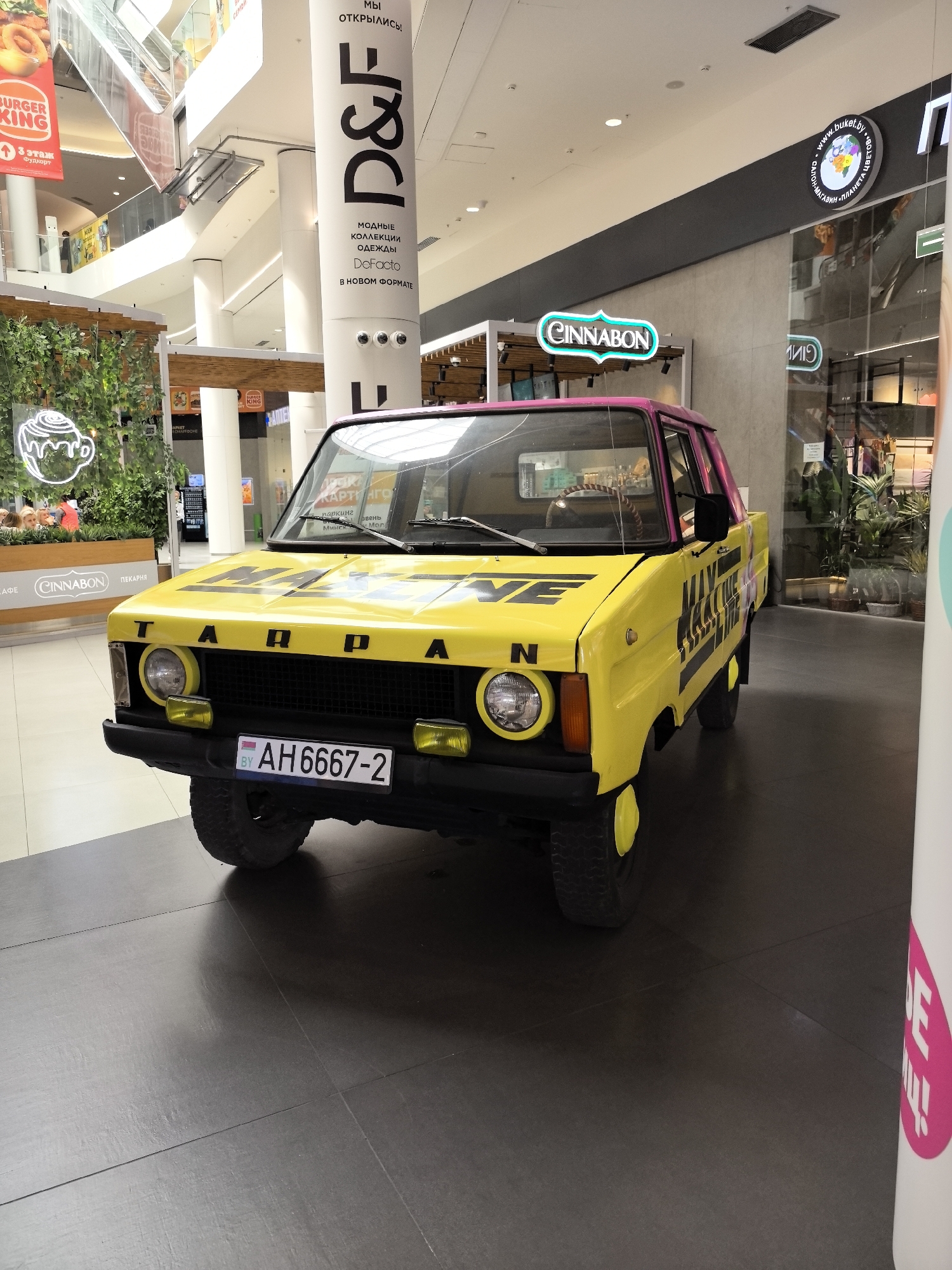 A bright yellow, vintage pickup truck is parked on a dark tiled floor inside a modern shopping mall. The truck has a pink roof and a license plate that reads "BY AH6667-2". Black lettering on the hood of the truck reads "TARPAN". On the side of the truck, there is black lettering that appears to be a logo or advertisement. The truck is the main subject of the image, and it is positioned in the center. In the background, there are various shops and escalators, typical of a shopping mall environment. A large white pillar with black lettering and a Cinnabon sign are also visible. The lighting is bright, suggesting the interior of a well-lit public space.