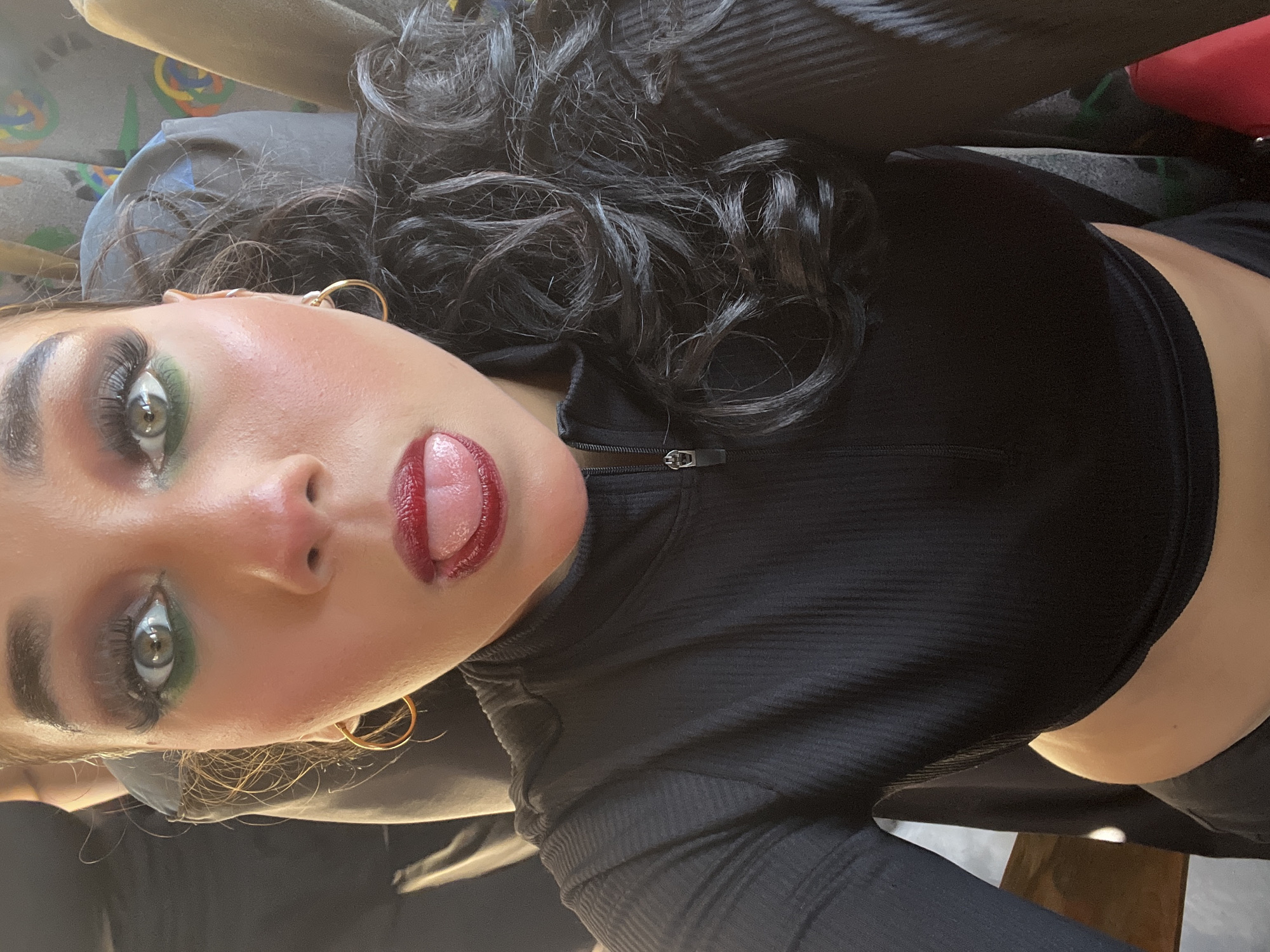 A close-up, vertically oriented selfie captures a young woman posing playfully. She has long, dark, curly hair and striking makeup, including vibrant green eyeshadow, winged eyeliner, prominent eyelashes, rosy blush, and glossy red lipstick. Her tongue is slightly extended. She is wearing a black, ribbed, long-sleeved crop top with a silver quarter-zip detail. Gold hoop earrings adorn her ears.
The background, mostly out of focus, reveals the upholstery of what appears to be a seat, possibly in a vehicle like a bus or car, characterized by a grey fabric with abstract, colorful patterns. The image is taken from a low angle, looking upwards at the woman. Her expression conveys confidence and a playful mood. The scene is set in an interior space in Porlamar, Venezuela. There is no visible text in the image.