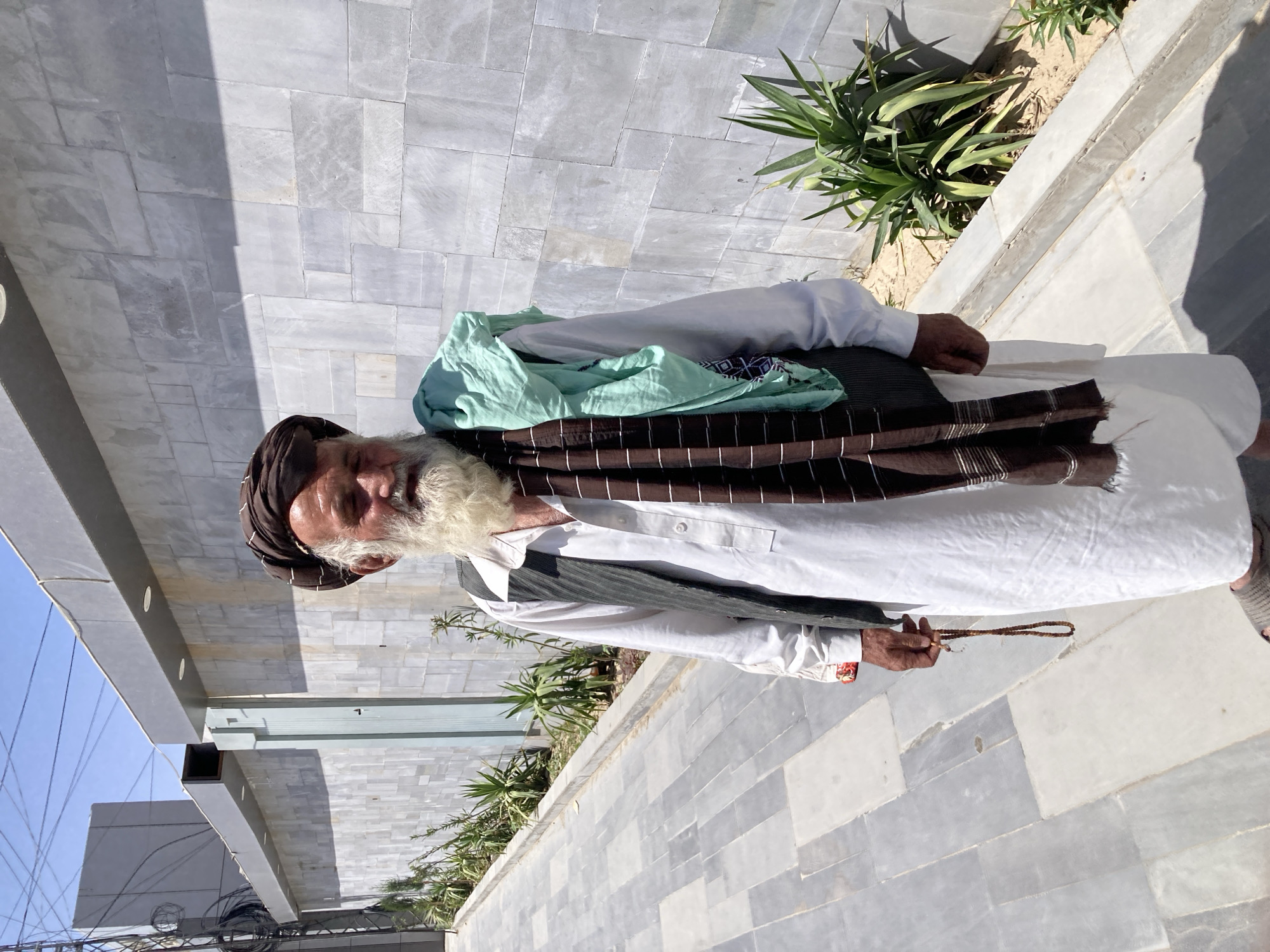 A mature man stands outdoors, possibly in the city of Quetta City Tehsil, Pakistan. He is wearing traditional attire: a brown turban, white shirt, dark vest, brown and white striped shawl, and light green scarf. He holds a string of beads in his right hand. The background includes a wall constructed of gray stone, plants in a planter, a gray building, and a blue sky.