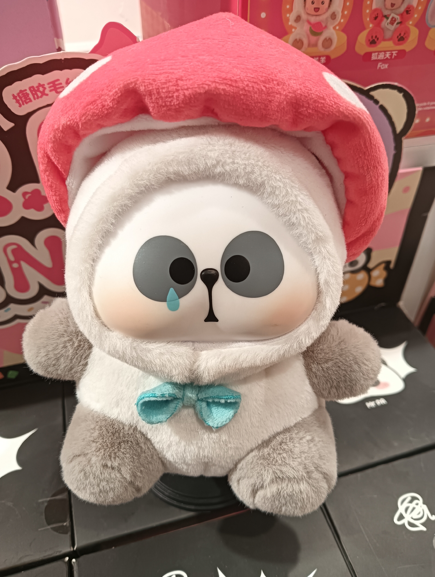A close-up, eye-level, medium shot captures a stuffed toy with a panda-like face and a red mushroom-shaped hood. The toy is predominantly white and grey, with large, circular grey eyes featuring black pupils and a small, light blue tear drop on its right cheek. It wears a light blue bowtie. The toy is seated on a black surface with white graphic designs, possibly part of a display or shelf. In the background, blurred pink and white elements suggest retail packaging or a shop environment. The image is well-lit, with the focus sharply on the stuffed toy. The scene does not contain any identifiable landmarks or elements that would place it specifically within Kediri City, Indonesia.