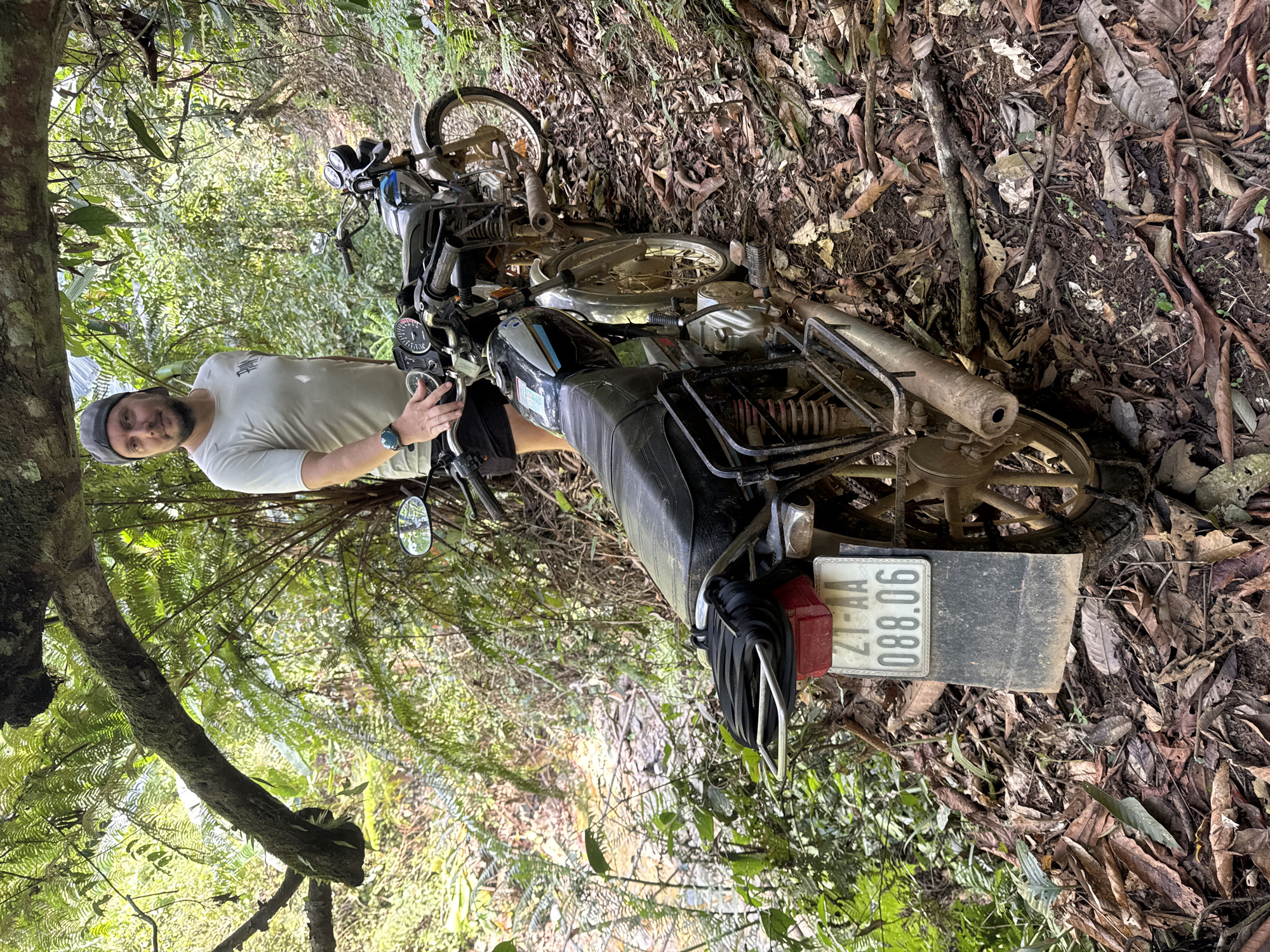 A man stands next to a fallen motorcycle in a wooded area. The motorcycle is lying on its side amid a ground covered in leaves and vegetation. The license plate on the motorcycle reads "21-AA 088.06". The location is Suối Giàng, Vietnam, as indicated by the license plate prefix. The man is wearing a light-colored t-shirt and a hat. He is holding onto the motorcycle. The environment is dense with foliage, including trees and ferns.