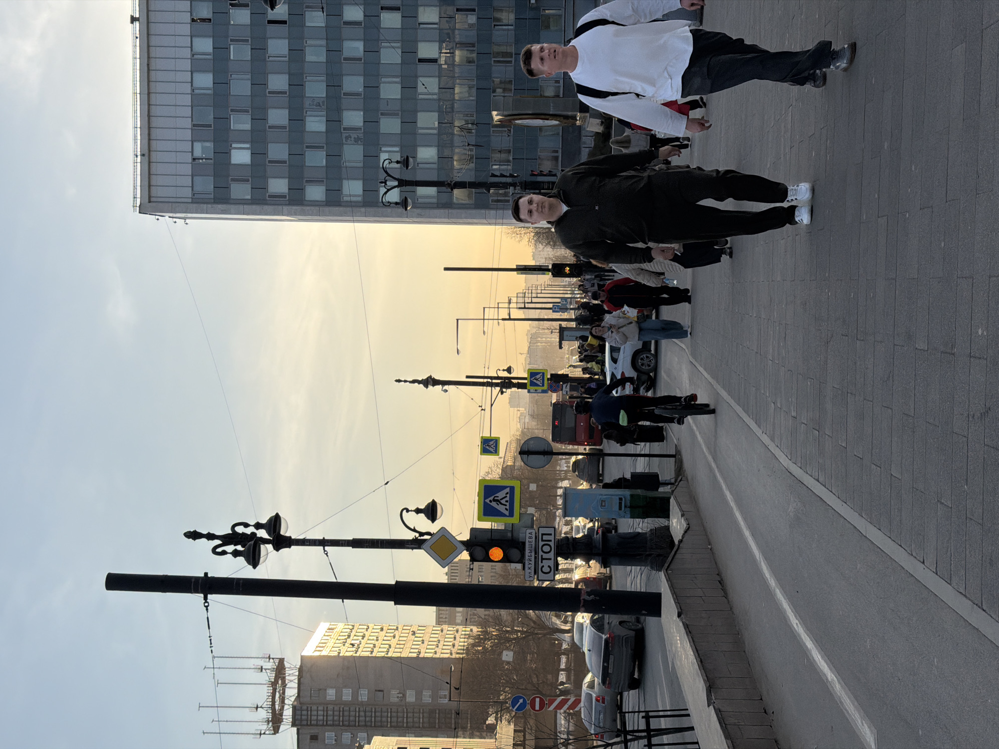 The image depicts a street scene in Perm, Russia. Two men are walking on the sidewalk, one wearing a white shirt and dark pants, the other in dark clothing. Behind them is a multi-story building with a grid-like window pattern. The street below is populated with pedestrians, cyclists, and vehicles, including cars and a bus. A traffic light is visible with an orange signal illuminated. There are numerous street signs lining the road. The sky is light, suggesting it is daytime. The overall scene appears to be a normal urban environment.