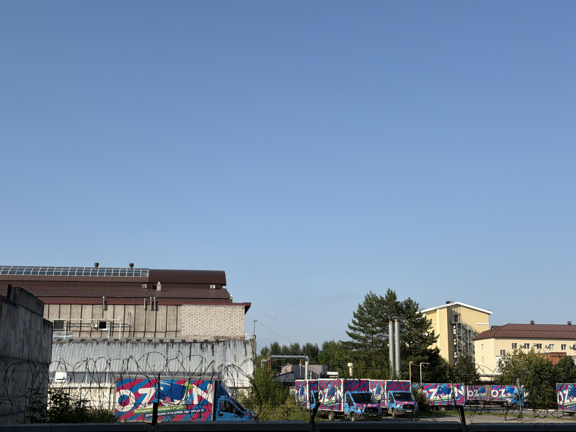 The image depicts an industrial area on a clear, sunny day with a bright blue sky. In the foreground, barbed wire fences are visible, with several blue vans decorated with colorful "OZ" branding parked behind them. The vans appear to be part of a delivery fleet. Behind the vans, industrial buildings with various architectural styles are present. One building has a brown, pitched roof with what appears to be solar panels. Another building is constructed with white brick. Further in the background, a yellow building with an external metal staircase and a brown-roofed building with multiple windows can be seen. Lush green trees are scattered throughout the scene, adding a touch of nature to the industrial landscape. The overall impression is one of a commercial or logistics hub. Based on the "OZ" branding on the vans, which is a well-known Russian online retailer, this scene is likely located in Tyumen, Russia, where "OZON" has a significant presence.