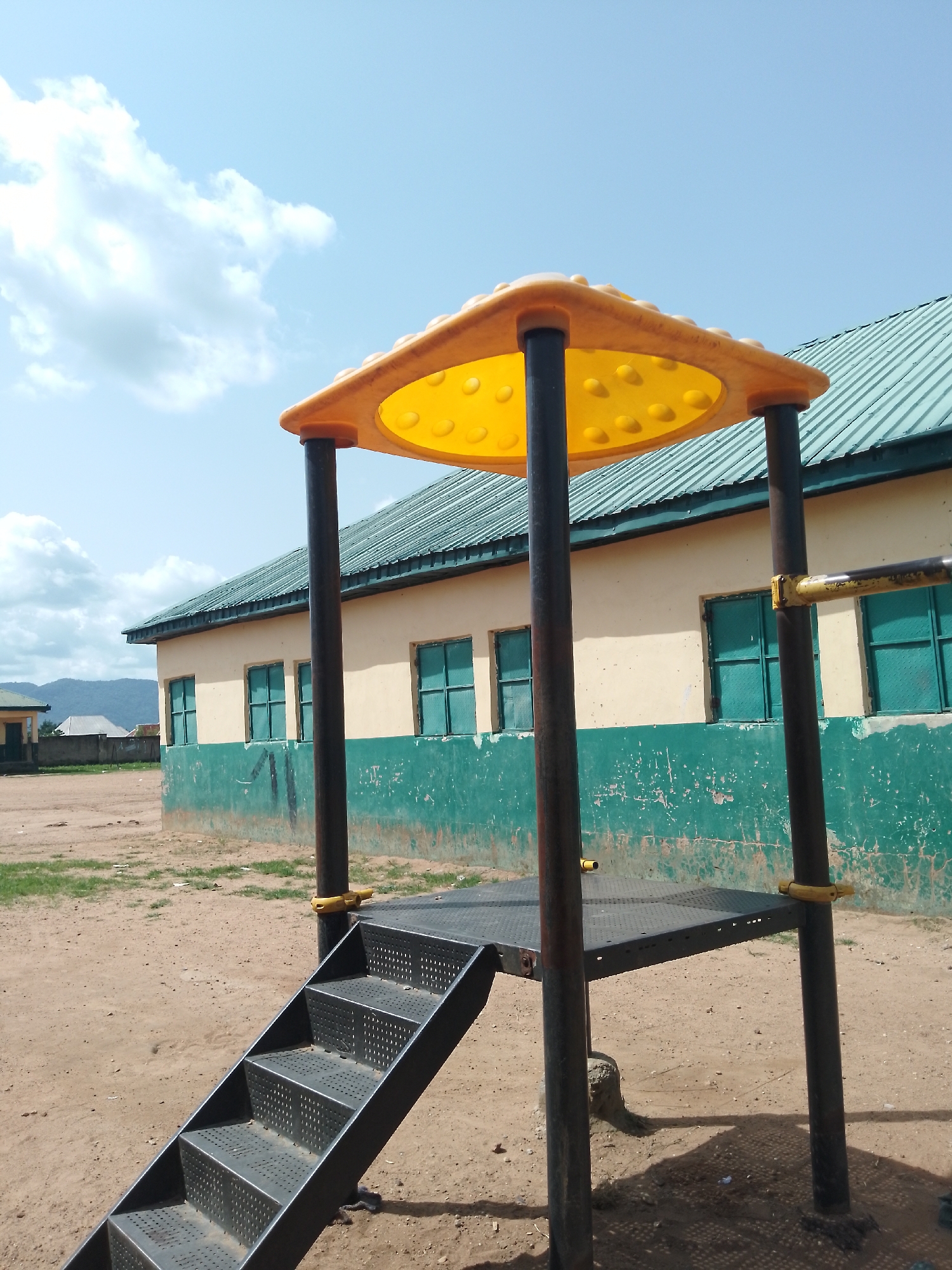 The image depicts a children's playground structure with a yellow roof and dark gray support poles, situated in the city of Serti, Nigeria. The playground is located in an outdoor, dusty area, likely part of a school or community center, given the presence of a building in the background. The building is a single-story structure painted in a light yellow and green color scheme. It features several windows with green bars. The roof of the building is a corrugated green metal. The sky above is bright blue with scattered white clouds. The ground is composed of dirt and sand with patches of dry grass. There are no people visible in the image. The playground structure itself includes a metal platform with perforations and a set of metal stairs.