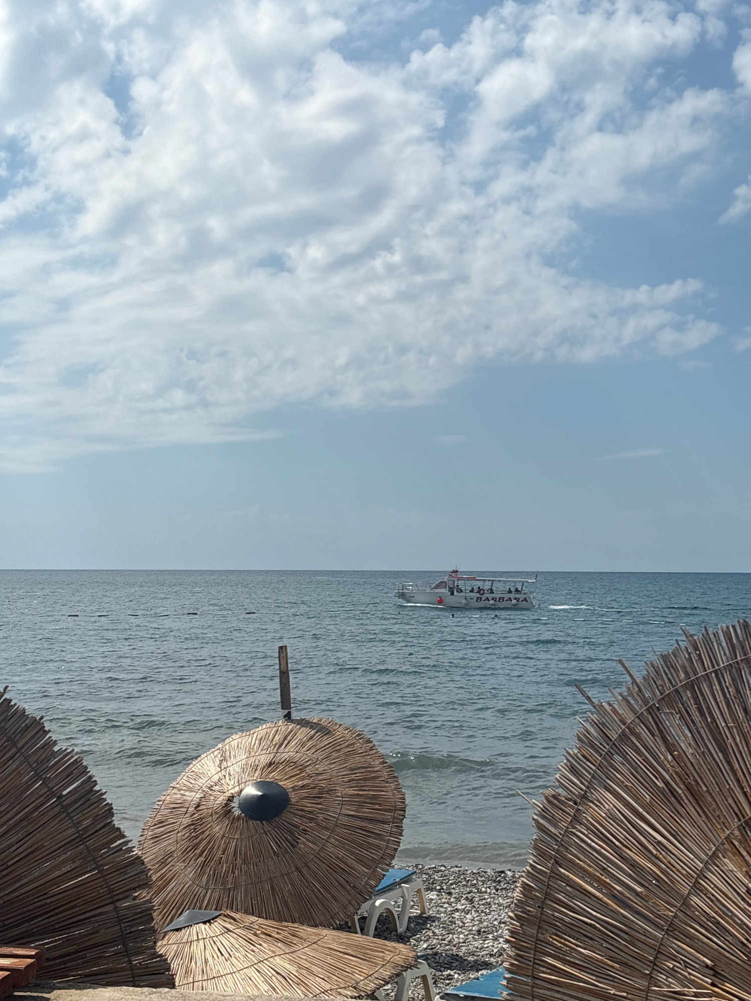 The image depicts a beach scene in Bar, Montenegro, under a partly cloudy sky. In the foreground, three thatched beach umbrellas are visible, two of which are fully in frame and one partially. One of the umbrellas has a black cap on top. Beyond the umbrellas, a pebbly shore leads to the calm, blue sea. A small white boat with people on board is sailing on the water in the mid-ground, with the word "BARBARA" visible on its side. The horizon line is clearly defined, separating the sea from the sky. The overall impression is one of a serene beach on a sunny day.