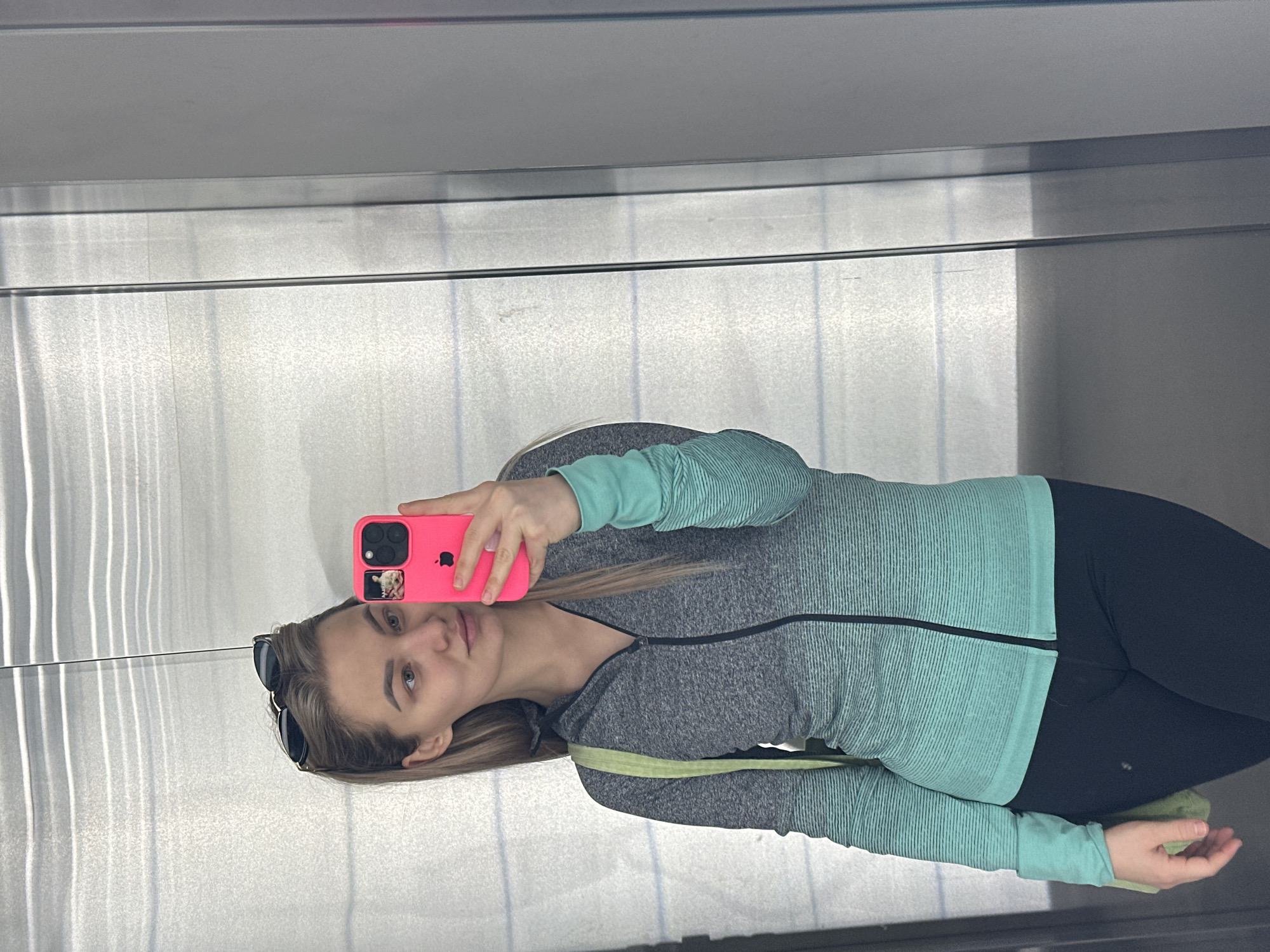 A woman is taking a selfie in an elevator. She is wearing a two-tone grey and light blue zipped athletic jacket and black leggings. She is holding a pink phone case with a sticker on it. Sunglasses are placed in her hair. The elevator walls are made of brushed metal. The location is unknown and there is no definitive evidence to indicate Budva, Montenegro.