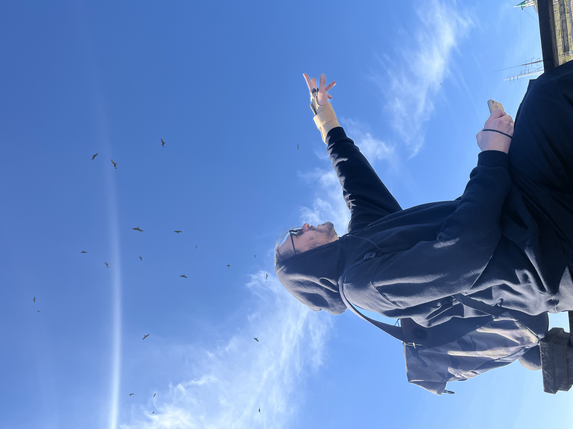 A low-angle shot depicts a man sitting on a ledge against a bright blue sky with scattered clouds and flying birds. The man wears a black hooded sweatshirt, black pants, and glasses. He has a beard and his right arm is raised with his fingers extended. He holds a phone in his left hand. A gray backpack sits beside him on the ledge. The location is Krasnoyarsk, Russia, based on the presence of overhead power lines and the unique architectural style of the ledge.