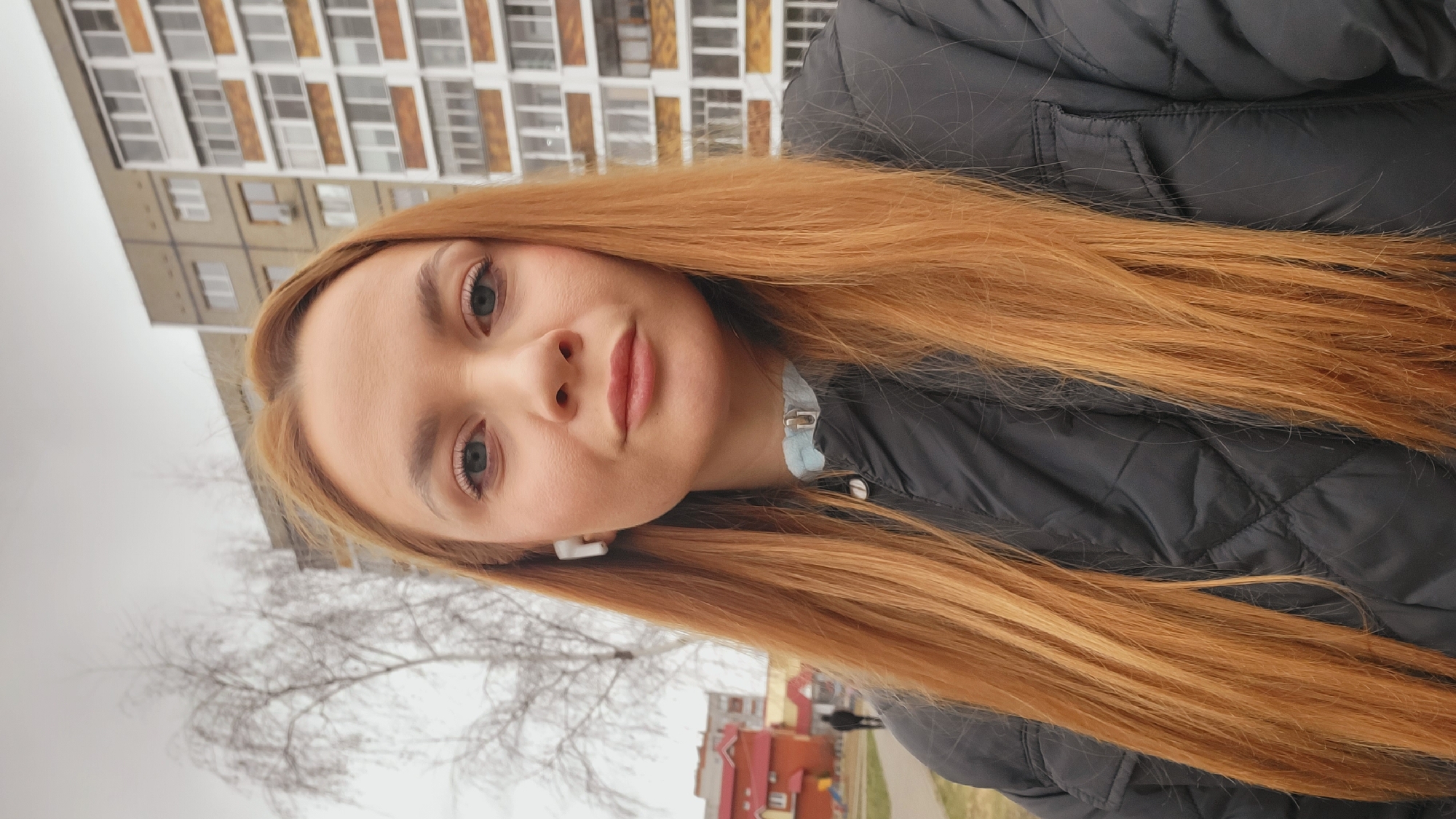 A close-up, chest-up portrait features a young woman outdoors in what appears to be an urban setting. She has long, straight, reddish-blonde hair that extends past her shoulders. Her light-colored eyes (possibly blue or grey) are directed towards the camera, and she displays a neutral to subtly soft expression. She is wearing a dark, possibly black or dark blue, quilted jacket, with a light blue detail visible around her neck or collar area. A white earbud is visible in her left ear.
The background consists of a large, multi-story residential building, likely an apartment block, characterized by its numerous windows and utilitarian architectural style, consistent with post-Soviet urban landscapes. Some windows have blinds or coverings. To the left, bare tree branches are visible against an overcast sky, suggesting the season is late autumn, winter, or early spring. Further in the distance, another building with a red roof can be seen. The overall scene indicates a daytime setting under cloudy weather conditions. There is no visible text in the image.
