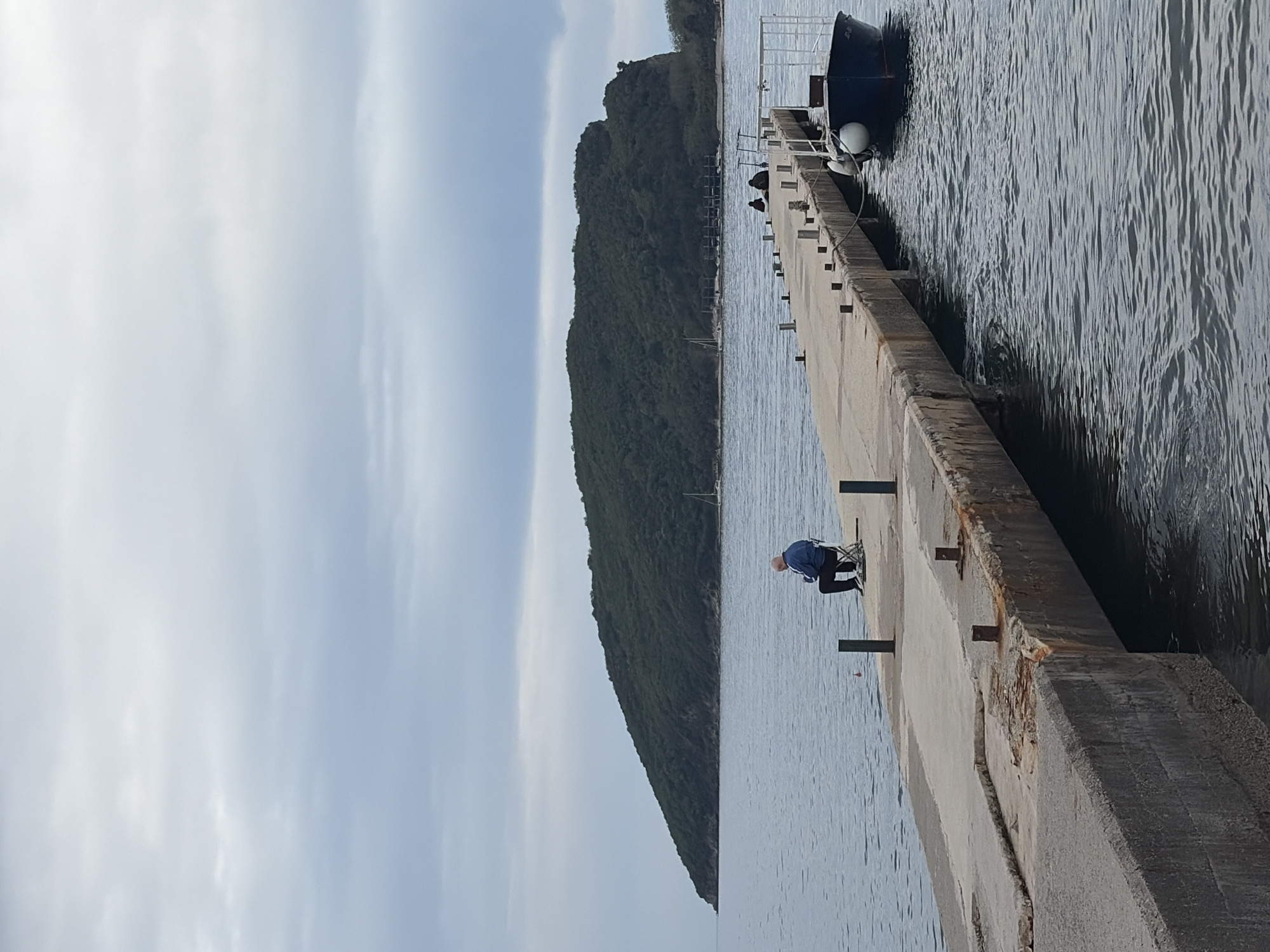 A pier in Budva, Montenegro, extends into the water. A person is seated on a chair near the edge of the pier, appearing to be fishing. Further along the pier, a small boat is docked. Trees line the adjacent land. The sky is overcast.