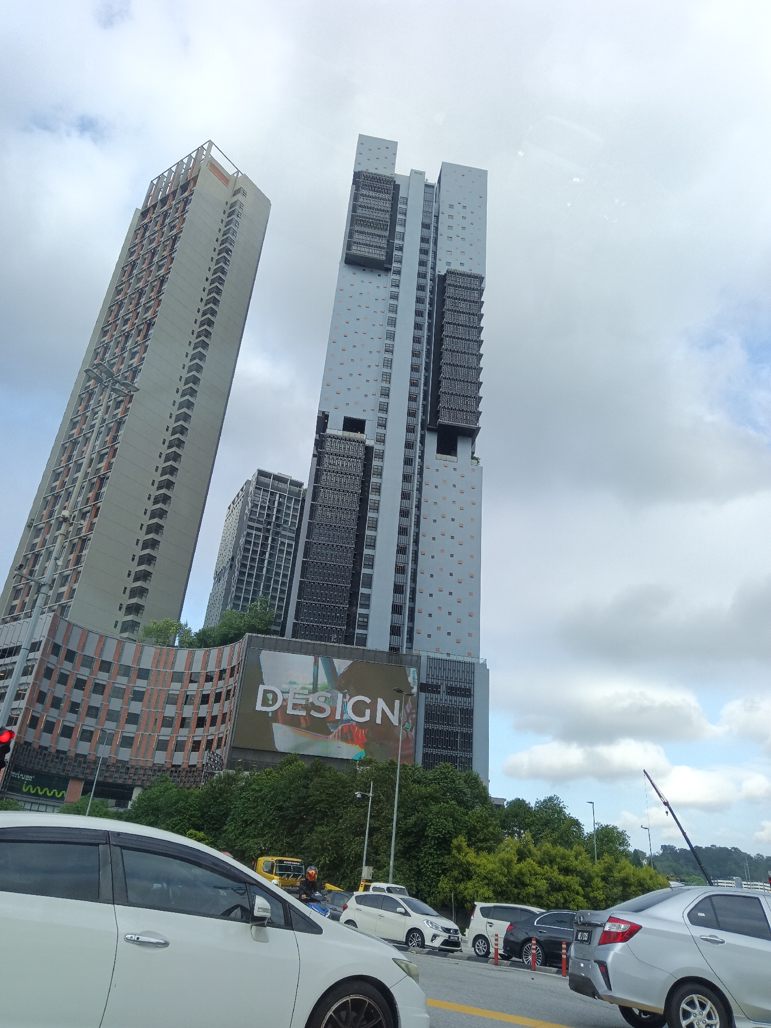 The image shows a street-level view of multiple modern skyscrapers under a cloudy sky in Kuala Lumpur, Malaysia. In the foreground, white and silver cars are visible in traffic, with a white sedan on the left taking up a significant portion of the frame. Behind the cars, a large billboard with the word "DESIGN" is prominently displayed on a curved building.
The skyscrapers dominate the background. The tallest, centrally located building is a light blue-gray structure with a distinctive pattern of small, square orange windows and dark, recessed sections. To its left is a much taller, slender building with a light gray facade and rows of orange-tinted windows. A shorter, darker building with a grid-like facade is situated between these two.
Green trees and foliage are visible behind the billboard and around the base of some of the buildings. A red traffic light is illuminated on the far left. A yellow truck and a person on a motorcycle are also present in the mid-ground, suggesting active traffic. The overall impression is one of urban development and modern architecture in a bustling city environment.