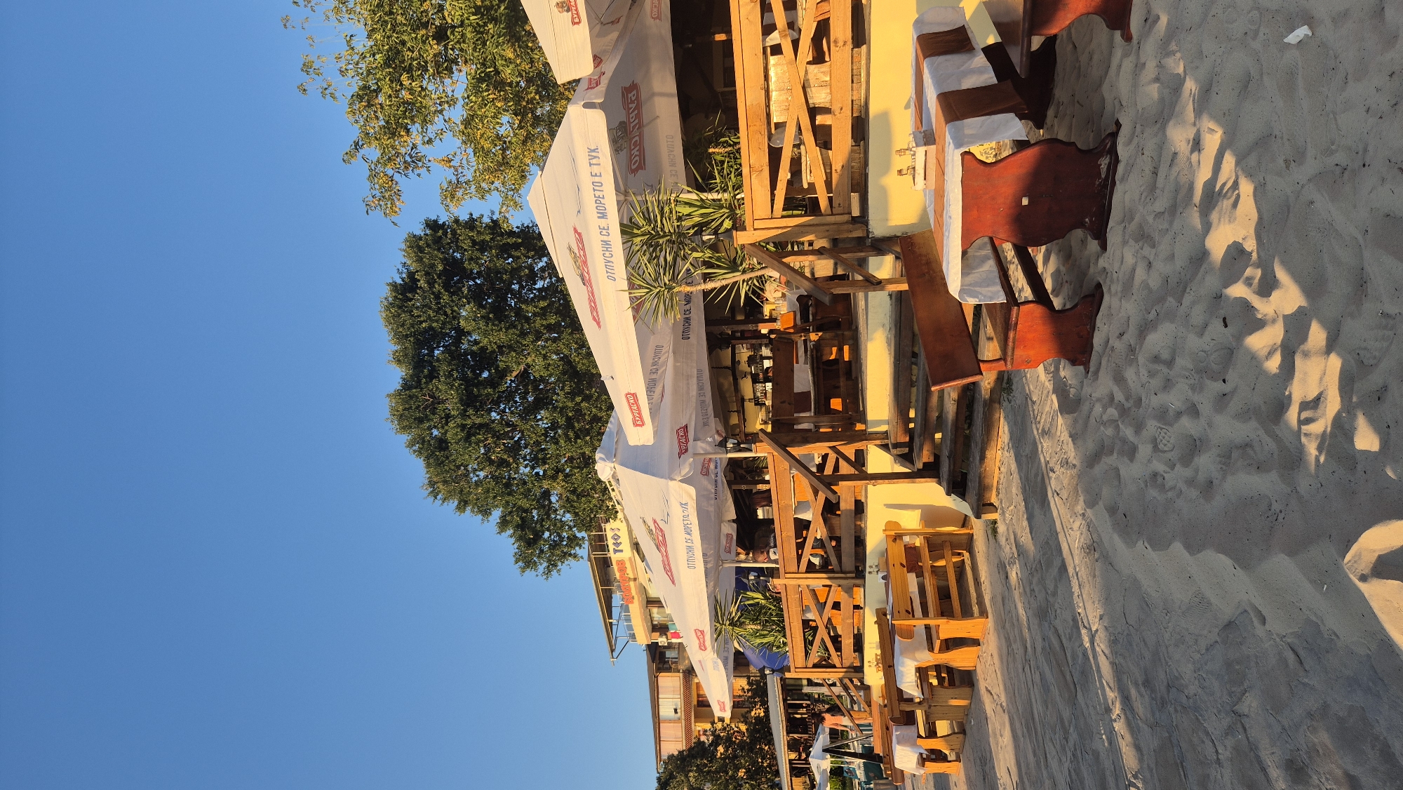 This is a landscape orientation, low-angle shot of an outdoor seating area on a sandy beach. The scene depicts several tables and chairs set up under large white umbrellas. The tables are covered with white tablecloths, some with brown runners, and appear to be set for dining. Wooden chairs, some with red cushions, are arranged around the tables. The sand is visible in the foreground and between the tables, with shadows cast from the umbrellas and a nearby building.
Behind the seating area, there are more structures with umbrellas and a glimpse of buildings. Trees, one large and leafy, are visible to the left, and the clear blue sky is above. The lighting suggests it is late afternoon or early evening, with a warm glow and long shadows.
Visible text on the umbrellas includes what appears to be Cyrillic lettering. One umbrella clearly shows "ОТИПУСНИ СЕ МОМЕТО Е ТУК" and another partially visible segment reads "ТУК". There are also logo elements that seem to be branded.
The setting is clearly a beachside establishment, likely a restaurant or cafe, in Kiten, Bulgaria, given the context. The overall mood is relaxed and inviting, suggesting a pleasant outdoor dining experience. There are no people clearly visible in the frame.