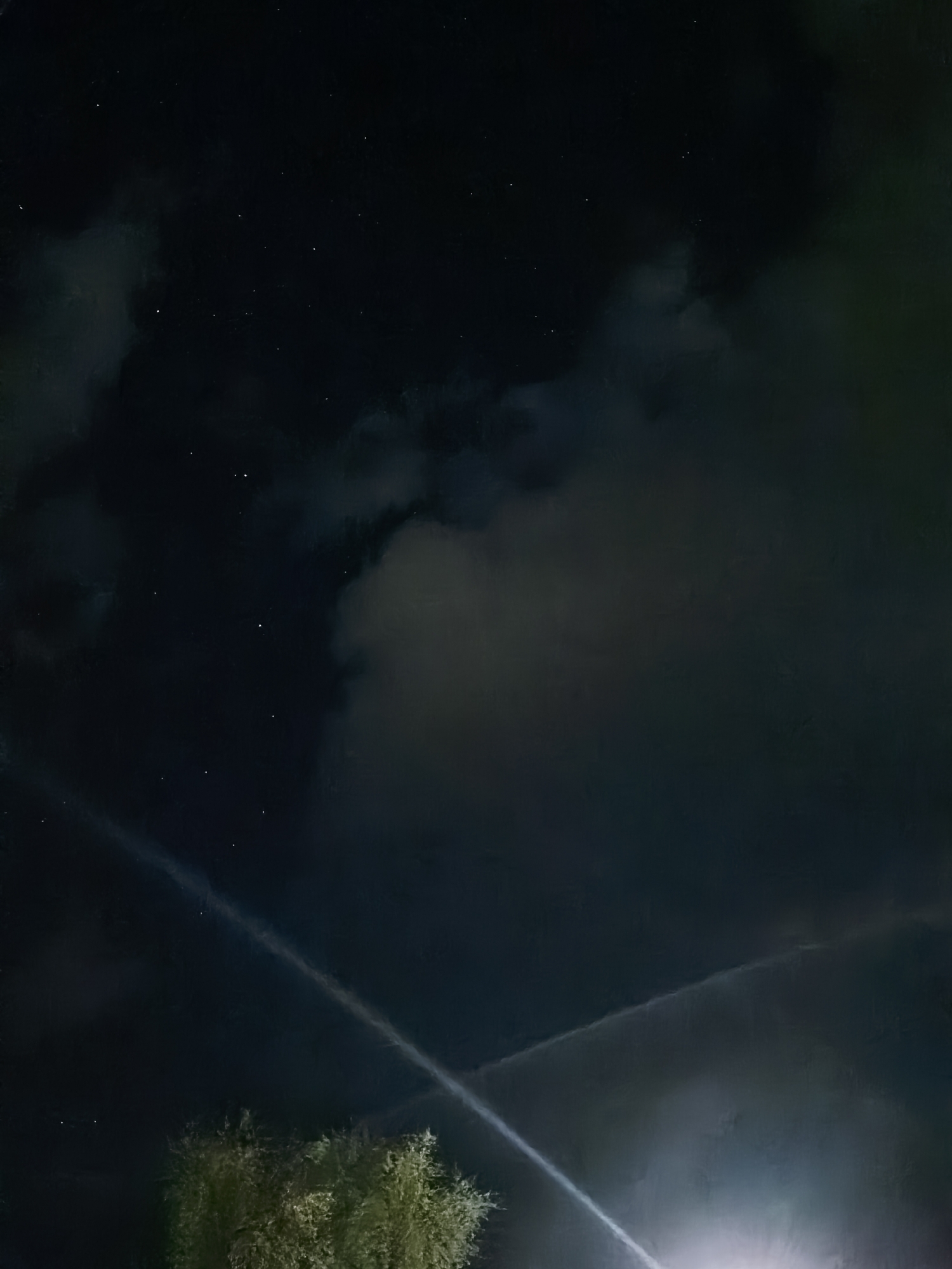 The image shows a nighttime sky with clouds and faint stars. A bright light source is visible at the bottom right corner, illuminating what appears to be the top of a tree. A faint line cuts diagonally across the sky from the bottom left to the upper right, possibly a contrail or light pollution. The location is in the city of Unknown, Unknown.