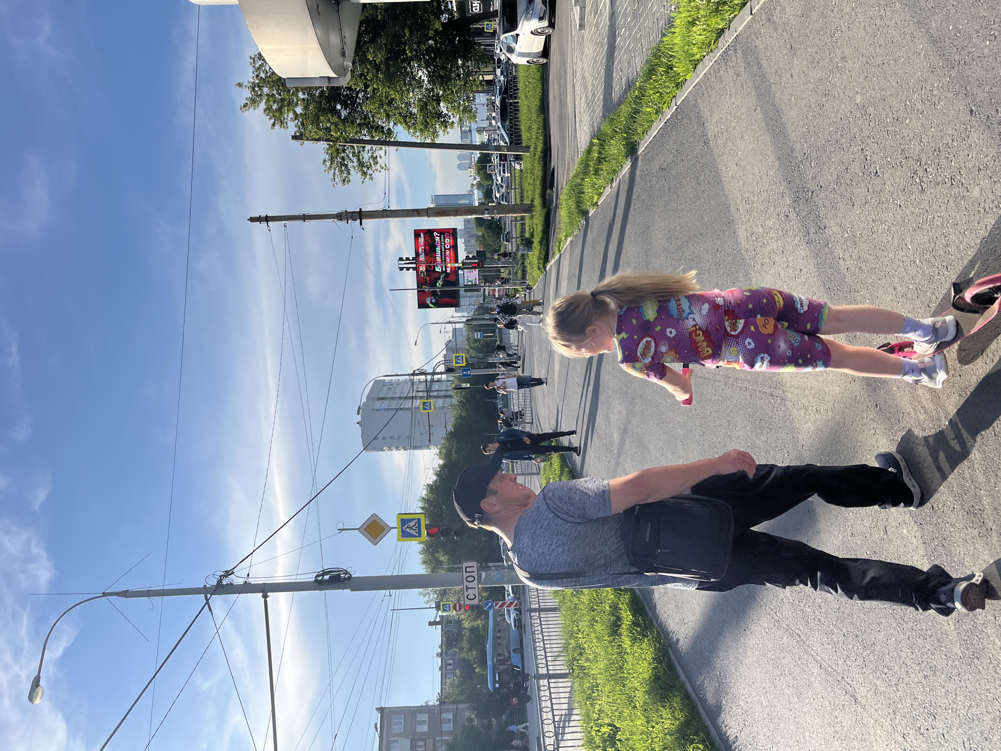 The image depicts a street scene in Yekaterinburg, Russia, on a sunny day with scattered clouds. In the foreground, a man wearing a grey t-shirt, black cap, and black pants, with a dark messenger bag slung across his body, walks away from the camera. Beside him, a young girl with blonde hair tied in a ponytail rides a pink scooter, also facing away from the camera. She is wearing a purple outfit with colorful comic book-style graphics and the word "BANG!" prominently displayed. They are on a paved pedestrian pathway adjacent to a grassy verge.
The background shows a typical urban environment with buildings of varying heights, a traffic light, street signs including a "STOP" sign, and a large digital billboard displaying advertisements. Electrical wires and poles are visible overhead, common in Russian cities. A blue bus, several cars, and other pedestrians are present on the street and sidewalks further down the road. Trees line the street, and the sunlight casts shadows on the pathway, indicating it is likely late afternoon. The overall atmosphere is one of casual city life.