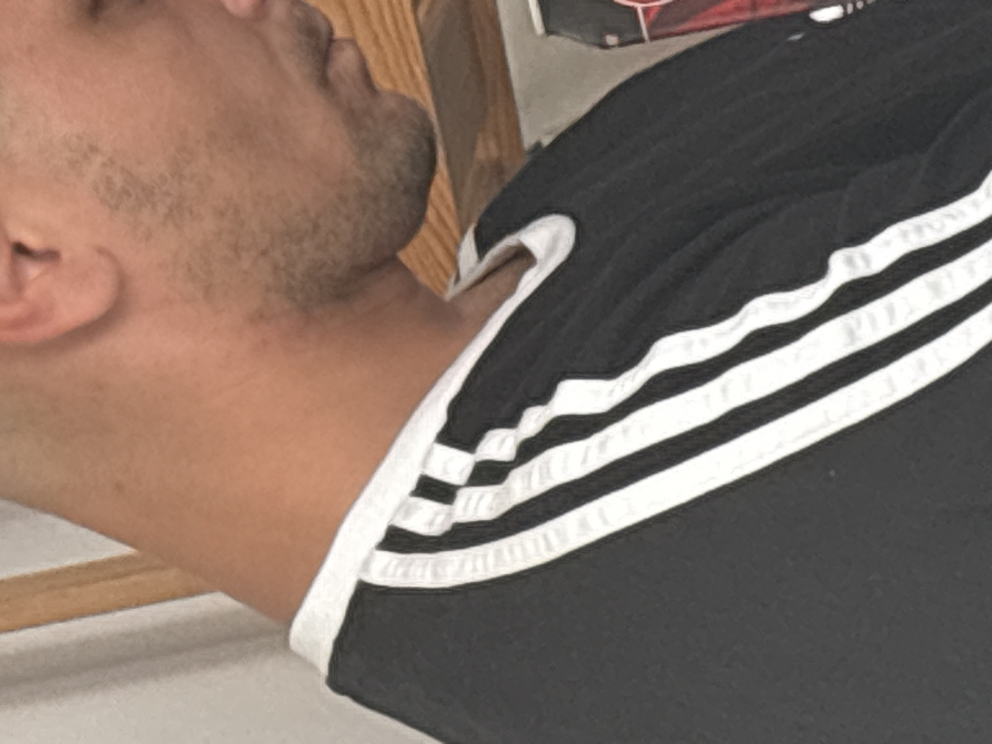 A close-up, waist-up, side profile of an adult male is captured in this indoor, natural light photograph. The man is wearing a black t-shirt with white stripes down the left sleeve, indicative of an Adidas brand garment. His right ear is visible, and he has stubble on his face. He is positioned in front of a wooden surface and a grey wall. No specific location in a city is identifiable from the image.