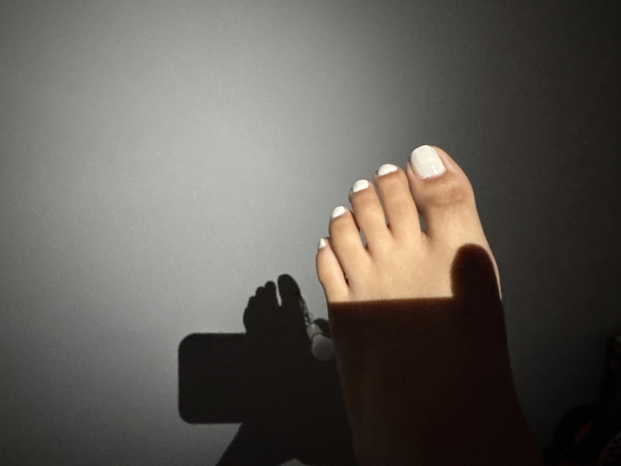 The image shows a person's foot with white nail polish against a gray wall. A shadow of a cell phone is cast on the wall behind the foot, indicating the phone was used to take the picture. The lighting suggests this scene is indoors, likely in a dimly lit room. There is no indication of the specific location within Madrid, Spain.