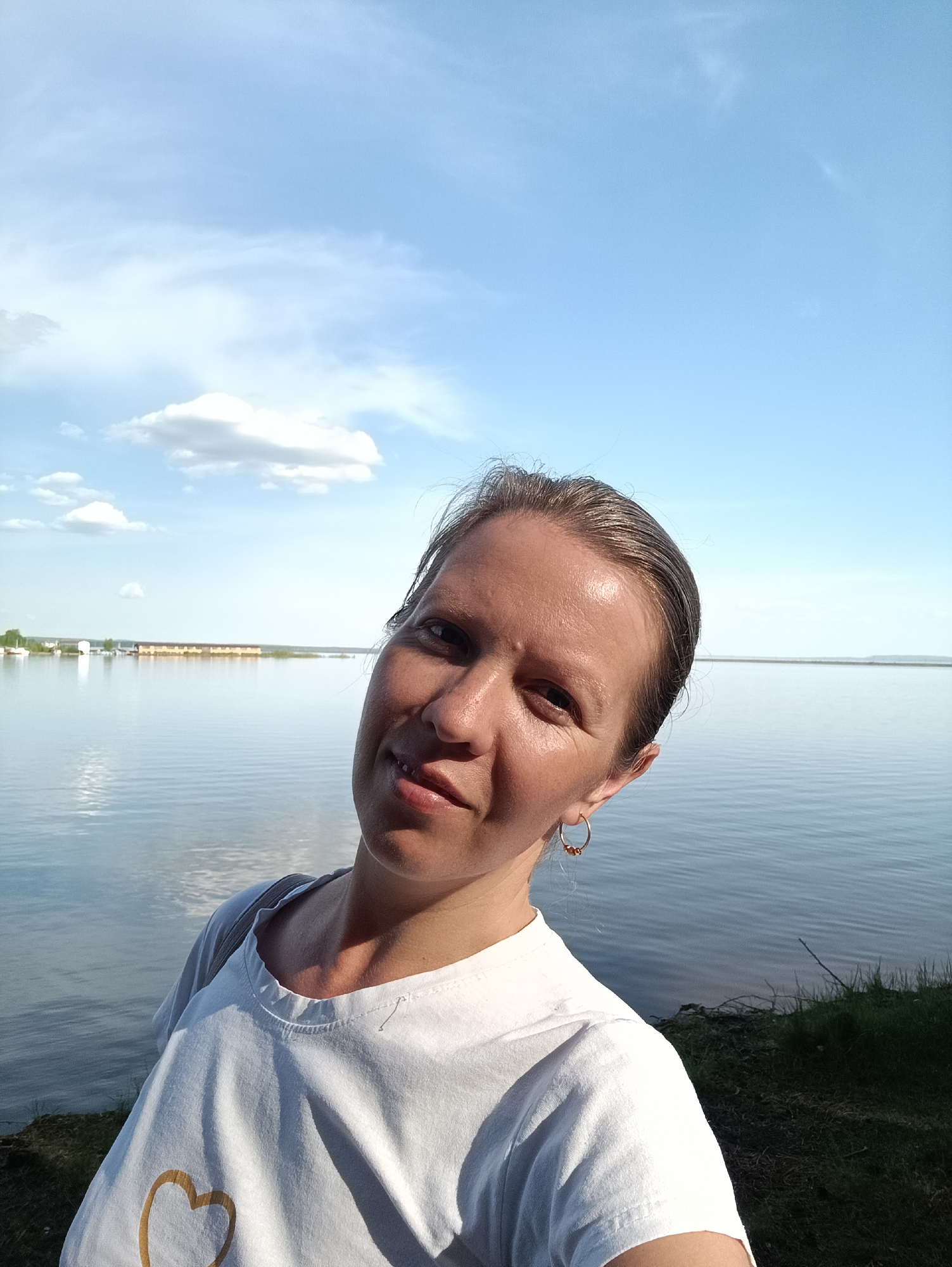 A woman is taking a selfie in front of a body of water under a blue sky with scattered clouds. She is wearing a white t-shirt with a heart design and a hoop earring. Behind her is a calm lake or river and a shoreline with green grass. In the distance, there are structures that may be buildings or docks. It is difficult to confirm this is in the city of Chaikovsky, Russia, based on the image.