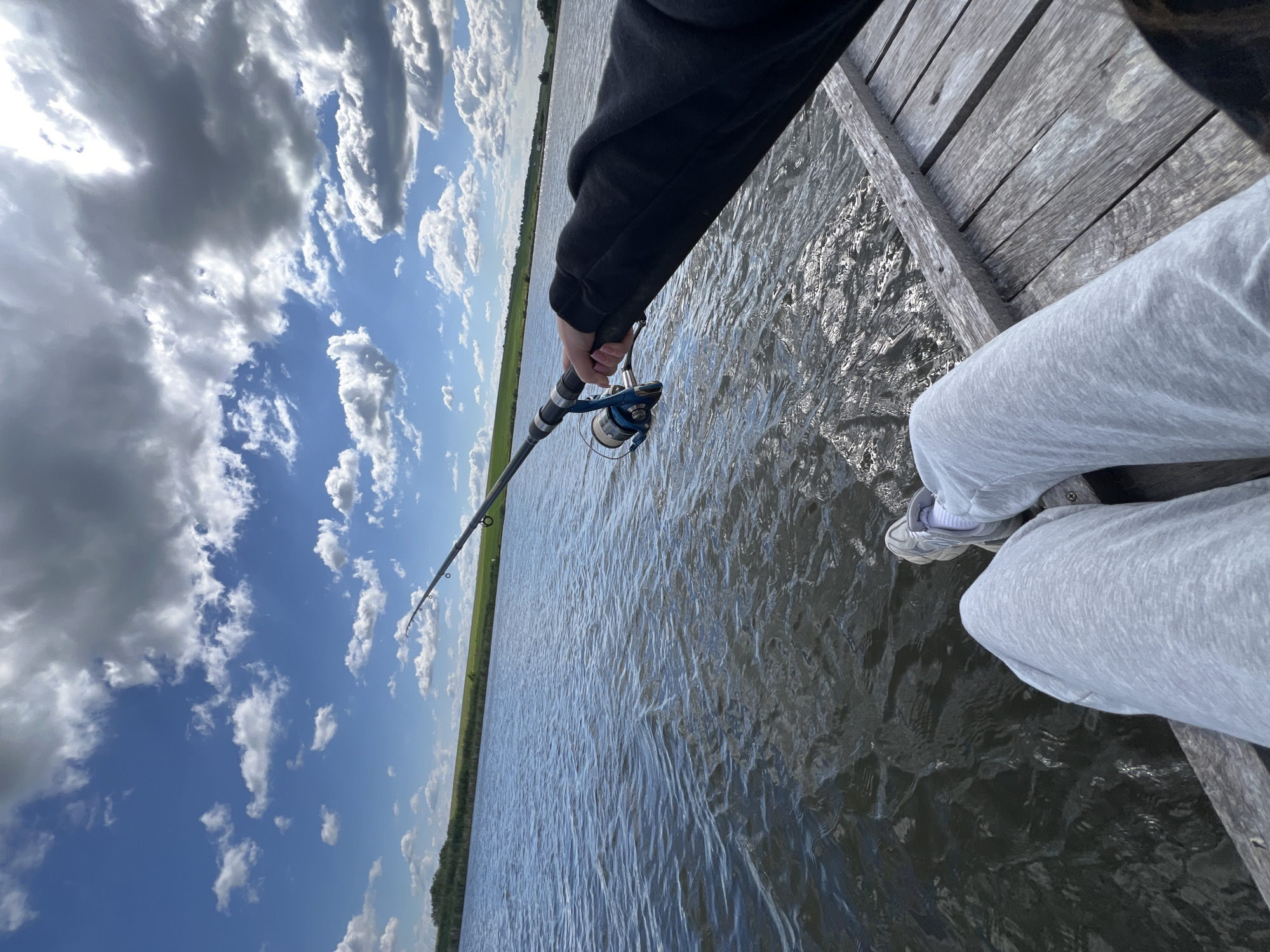 A person in grey sweatpants sits on a wooden dock and holds a fishing rod over the water. The sky is blue with puffy white clouds. A grassy shoreline is visible in the distance. The location of this scene is not possible to determine.