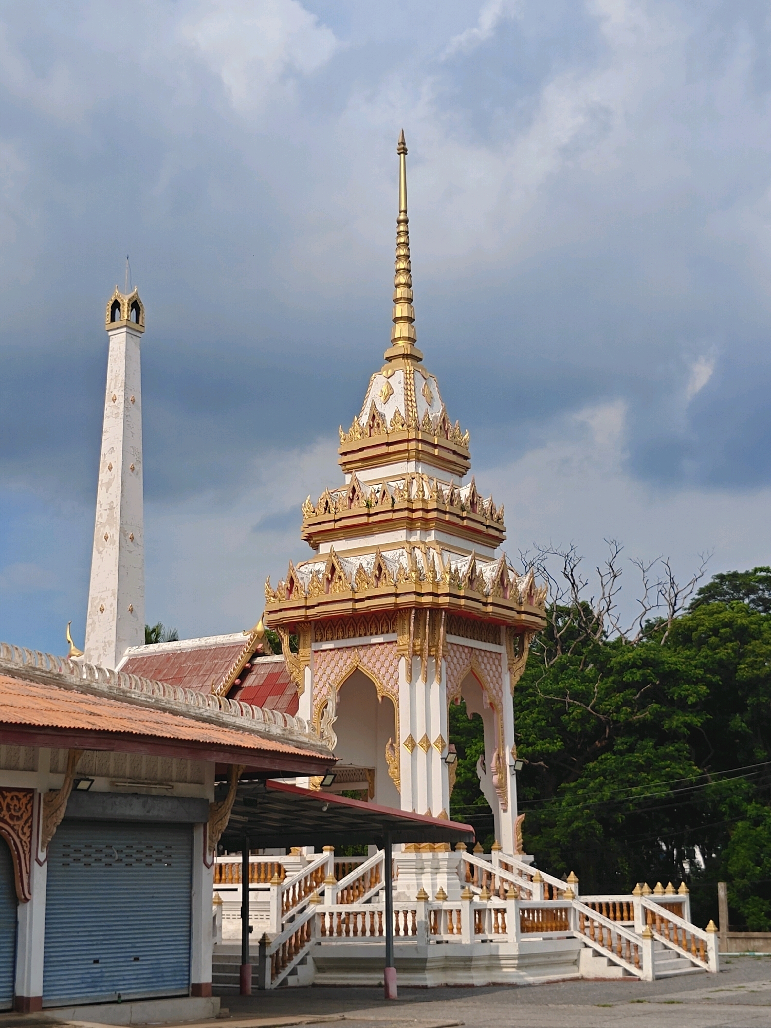 The image shows a crematorium structure with intricate white and gold detailing, including multiple tiers and a tall spire. To the left of the crematorium is a tall, white, rectangular chimney, also adorned with gold accents. In the foreground, a building with a red corrugated metal roof and blue roller shutters is visible. The structures are set against a backdrop of lush green trees and a cloudy sky. The scene appears to be located in a temple or a similar religious site within Phra Nakhon Si Ayutthaya City Municipality, Thailand, given the architectural style. There are no people or animals visible in the frame. The overall impression is one of a solemn and culturally significant location.