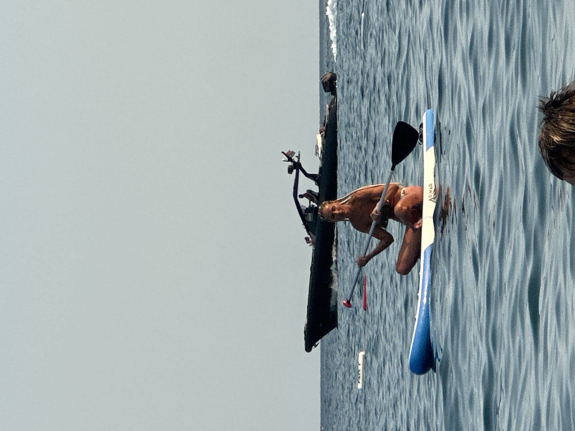A woman is paddleboarding on the water in front of a dark, sleek yacht. The woman is sitting on the paddleboard, holding a paddle with a black blade. She is wearing a bikini. The yacht is in the background, with a few people visible on it. The water is calm and blue, with small waves. The sky is clear and pale blue. The scene appears to be taking place on a sunny day. There is a hint of another person's head visible in the lower foreground of the image. The location is the coast of Boreti, Montenegro.