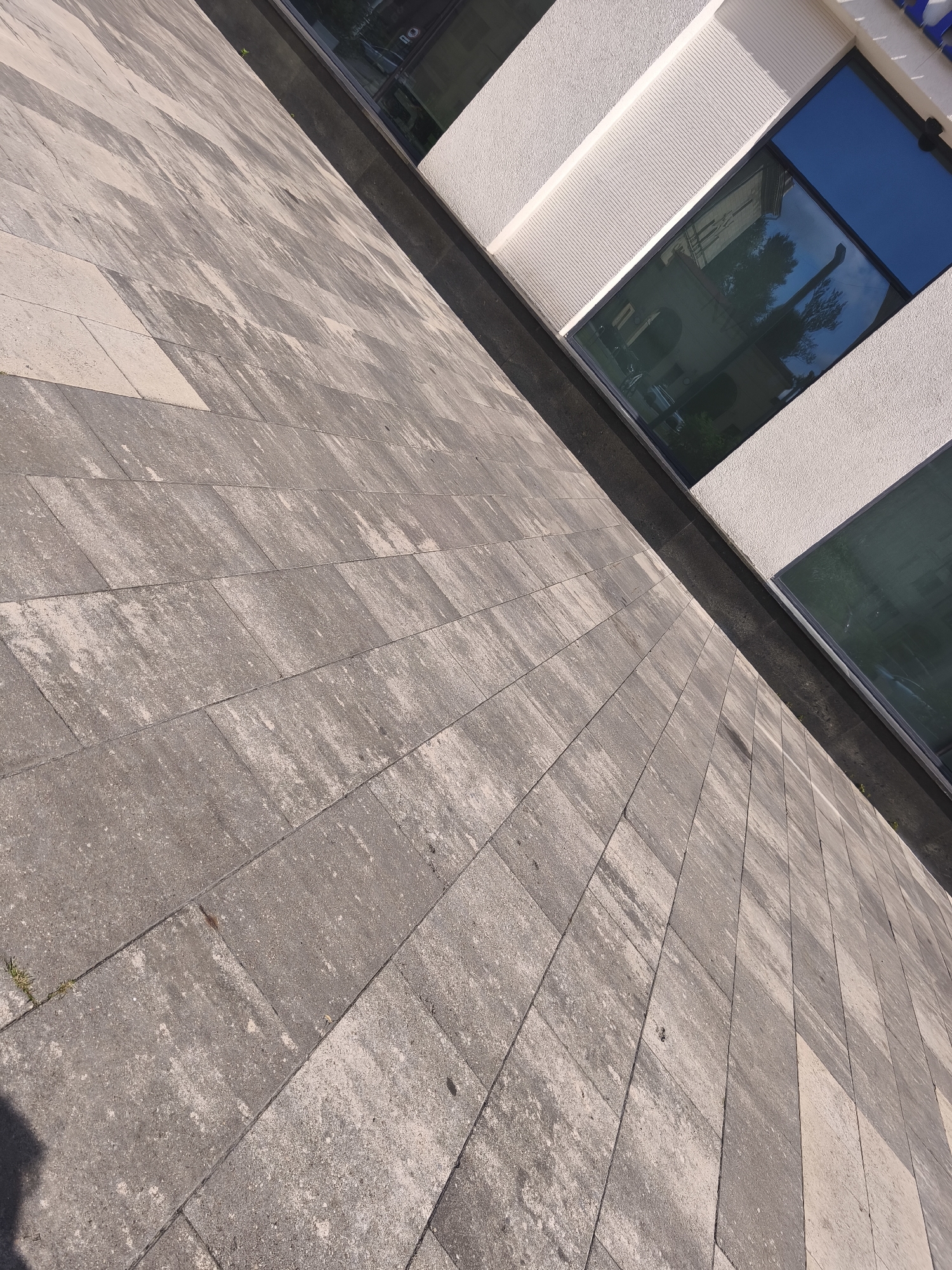 The image depicts an outdoor scene with a textured concrete pavement in the foreground, laid in rectangular stones with a stone-like appearance. The pavement stretches diagonally from the bottom left towards the top right. Above the pavement, a dark band, possibly a curb or trim, separates it from the building facade. The building features large, reflective windows that mirror the surrounding environment, including trees and a building facade. The building itself has a light-colored, textured exterior wall with horizontal grooved accents above the windows. The lighting suggests it is daytime, with shadows visible on the pavement. There are no people or distinct actions occurring in the image. The scene appears to be a public urban space, likely a plaza or sidewalk adjacent to a modern building. Based on the available context from the dataset, this image is located in the city of Minsk, Belarus.