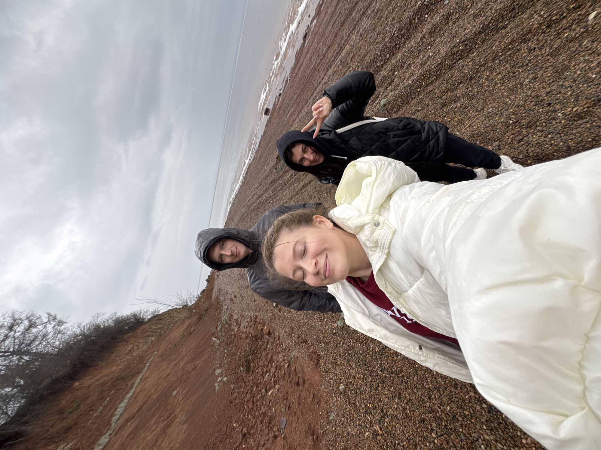 Three individuals are captured in a photograph taken outdoors. The setting appears to be a beach with a body of water, potentially a river or lake, visible in the background. The sky is overcast, suggesting a cloudy day.
The first individual, positioned closest to the camera, is wearing a white hooded jacket and appears to have their eyes closed. The second person, located behind and to the left of the first, is wearing a gray hooded jacket. The third person, situated further back and to the right, is dressed in a black quilted jacket and is holding up a peace sign with their fingers.
The ground is covered in pebbles or gravel. To the left of the group, there is a small cliff or embankment with sparse vegetation. The photograph appears to have been taken at an angle, giving the impression that the scene is tilted. Given the metadata provided, the location is the city of Chaikovsky, Russia.
