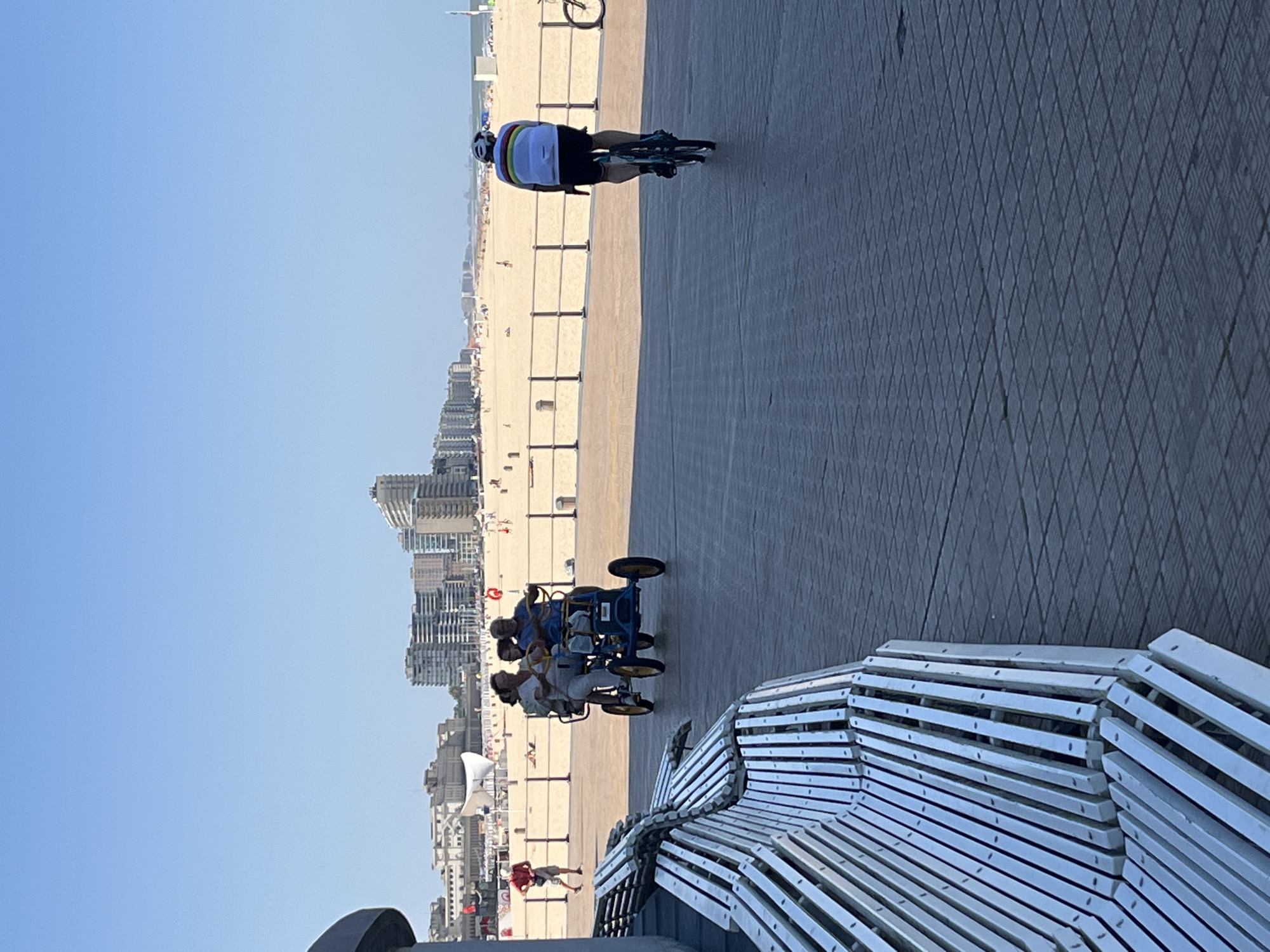 The image depicts a scene on the Ostend, Belgium, seawall promenade. In the foreground, a portion of a long, light-colored wooden bench with metal supports is visible on the left. The promenade surface is made of dark gray pavers arranged in a diamond pattern.
Further down the promenade, a multi-person pedal vehicle, colored blue with yellow wheels, is being ridden by three people. Two individuals are seated in the front, pedaling, and one person is seated behind them.
To the right of the pedal vehicle, a person on a bicycle is riding away from the camera. This cyclist is wearing a helmet, a white jersey with a horizontal striped pattern on the back, and dark shorts.
In the background, a sandy beach is visible, lined with beach umbrellas and people. Beyond the beach, a row of multi-story buildings forms the city skyline. The sky is a clear, bright blue. A person in a red shirt and shorts is walking on the promenade to the left of the pedal vehicle.