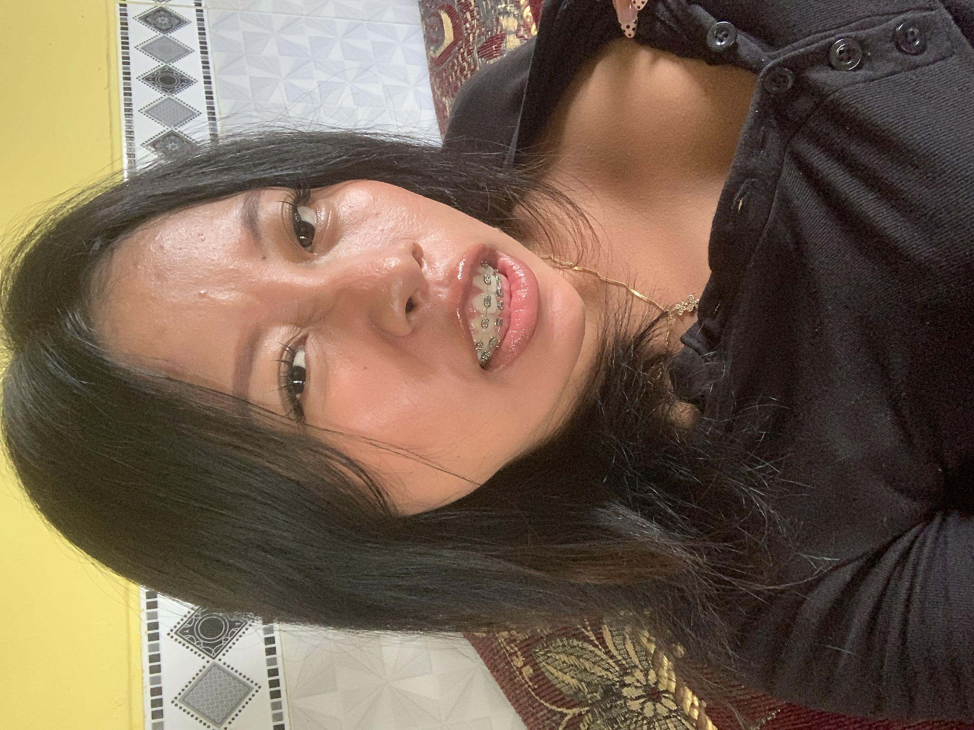 A woman with black hair and braces is looking into the camera. She is wearing a black shirt with buttons. The background consists of a yellow wall and a tiled wall. The scene appears to be indoors, likely in a residential setting in Jajag, Indonesia, as suggested by the style of the walls.